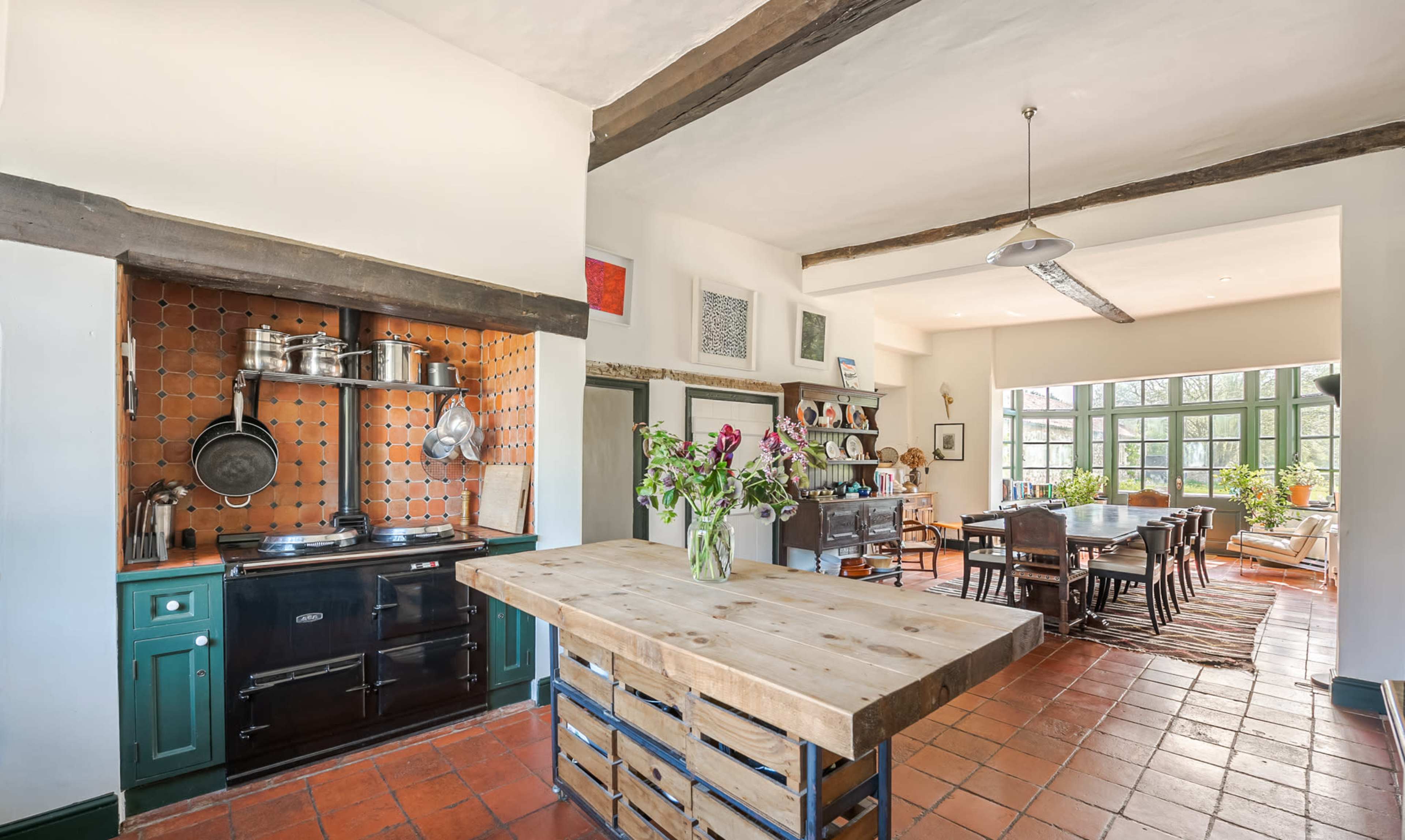 A spacious kitchen featuring a large central wooden island, a black stove with metal cookware, and a dining area with a long table and large windows.