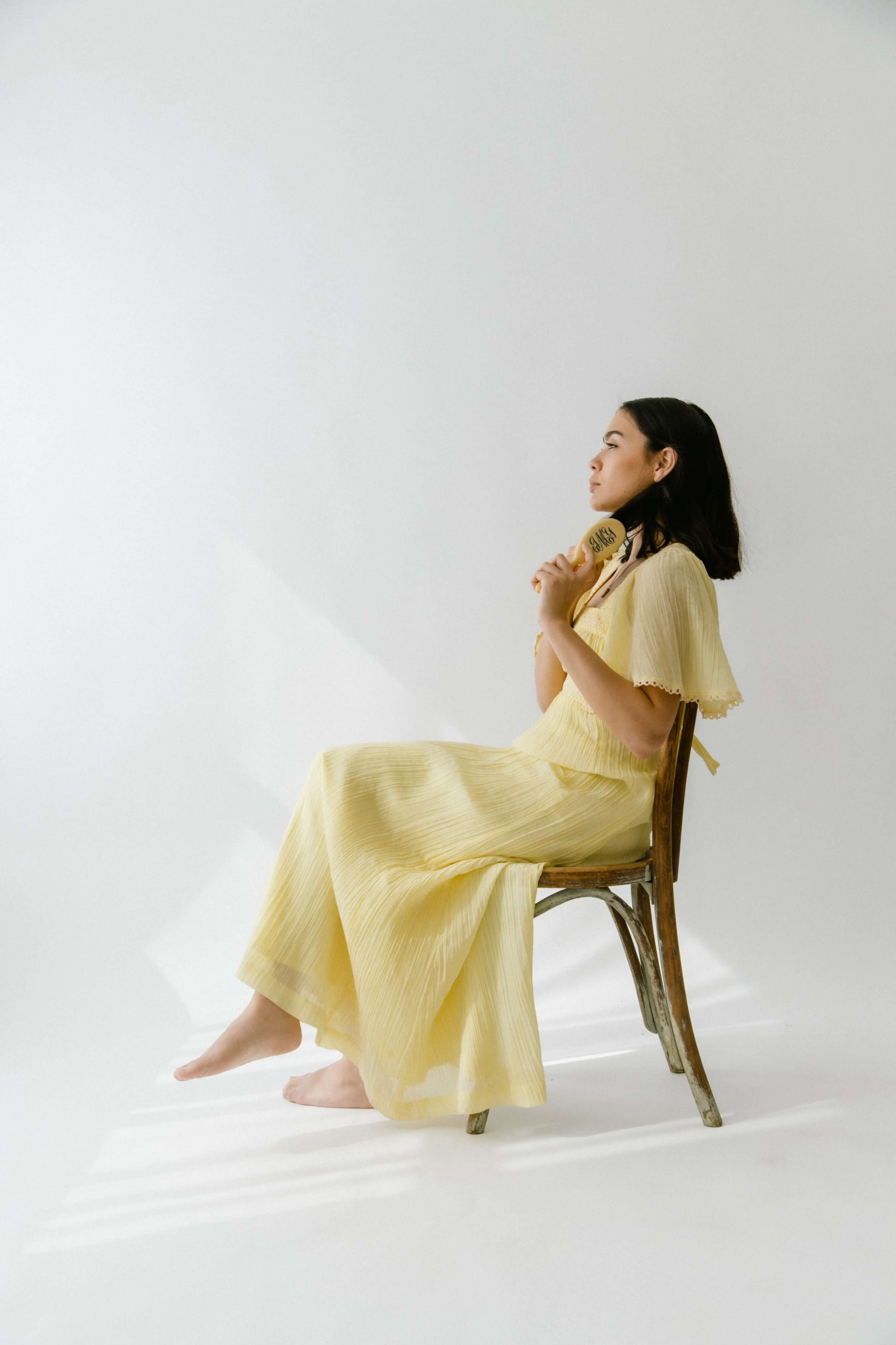 A woman in a yellow dress sits barefoot on a wooden chair, holding a decorative object while gazing to the side.