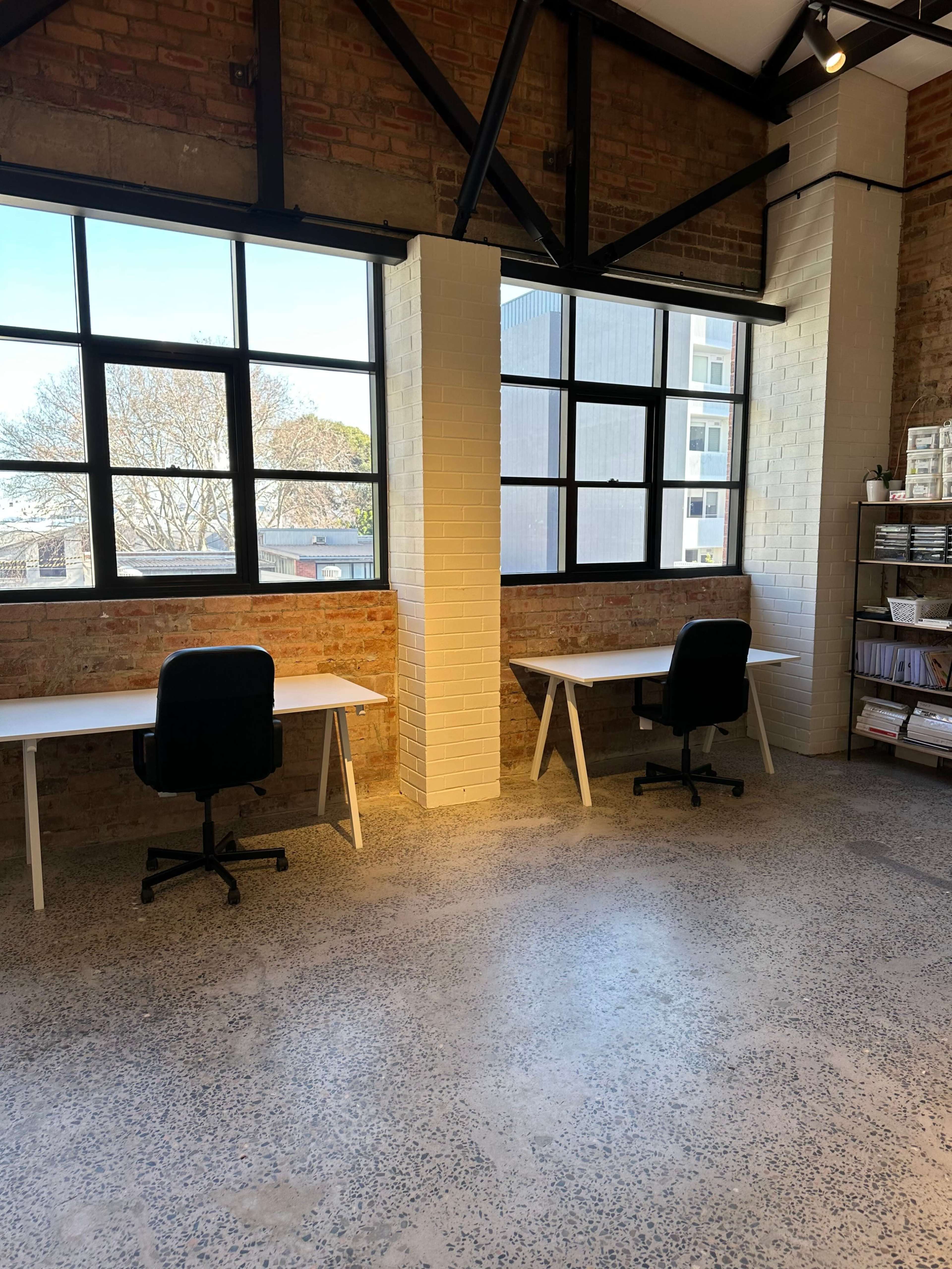 A spacious room with two white desks and black chairs positioned next to large windows, featuring brick walls and a polished concrete floor.