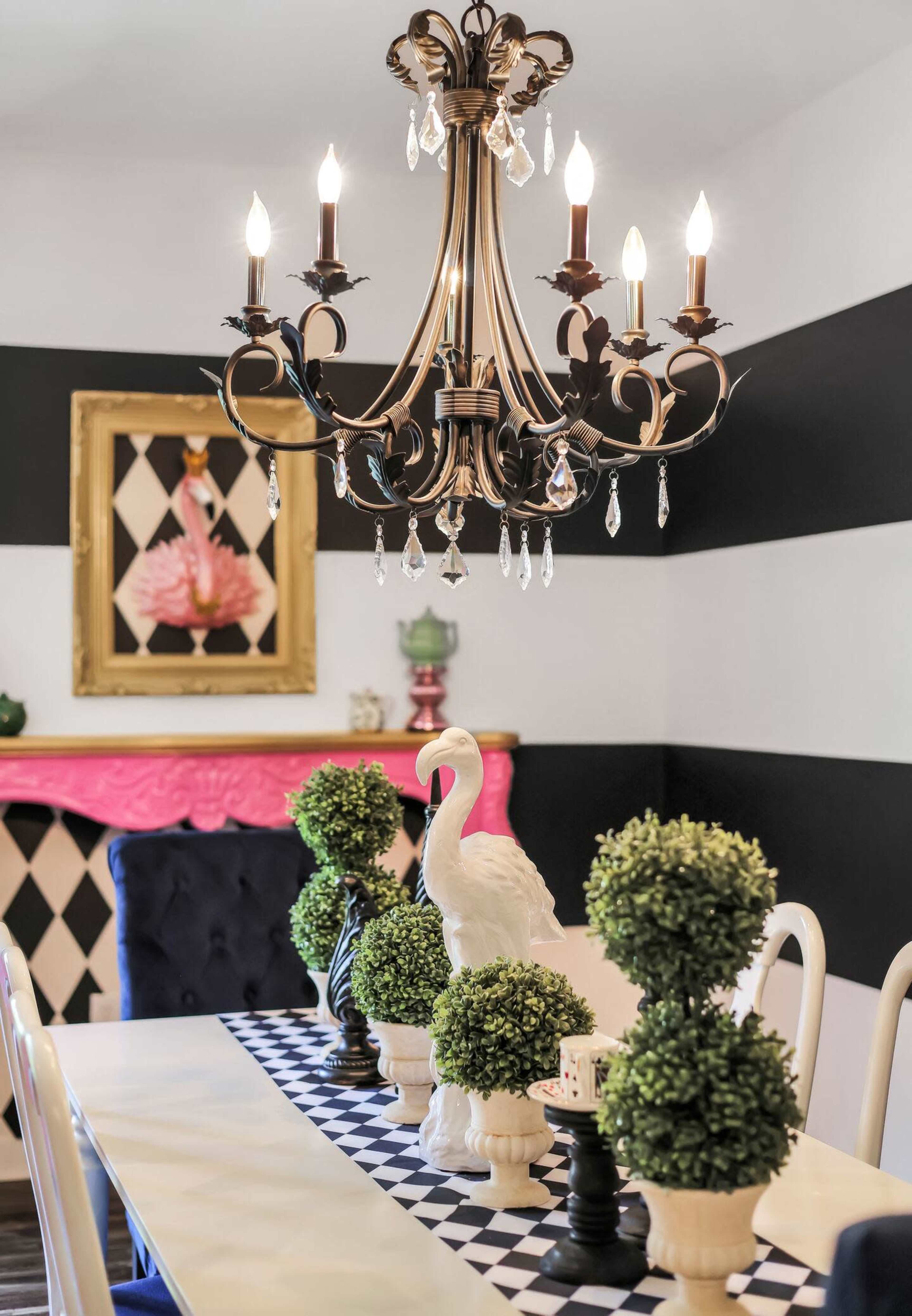 A dining room features a chandelier above a long table adorned with topiary plants, a decorative swan, and a black and white checkered table runner, with striped walls and a pink fireplace in the background.