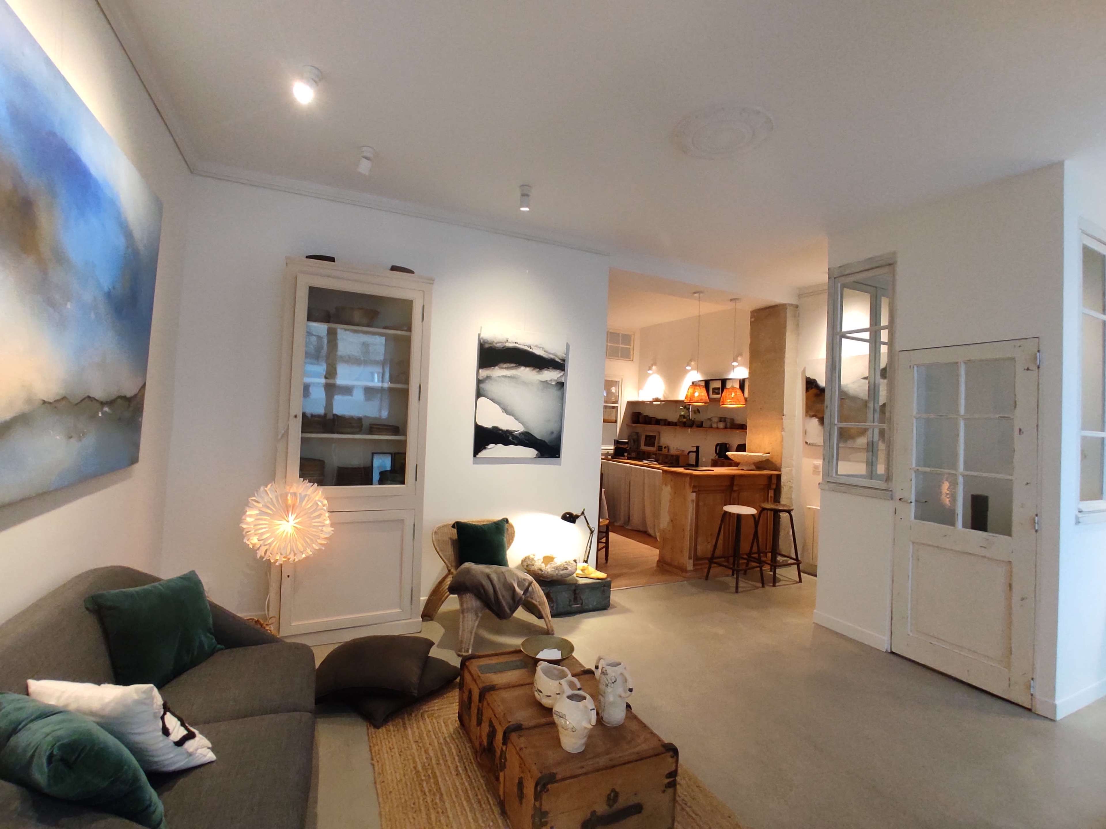 The image shows a modern living room with a grey sofa, a wooden coffee table, and a view into a kitchen area with bar stools and artwork on the walls.