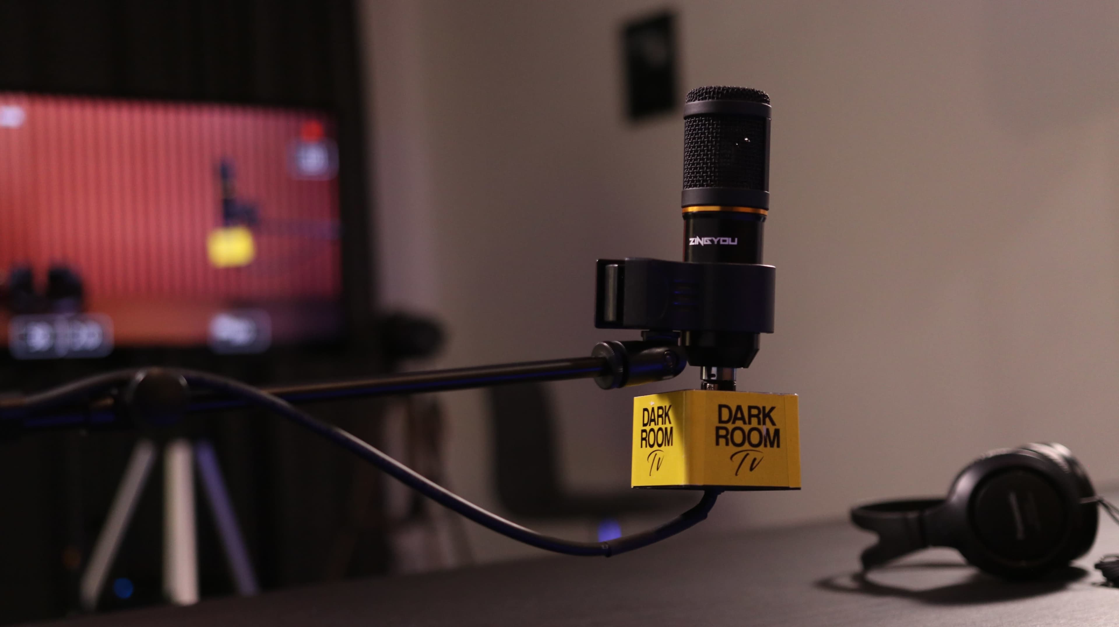 A microphone with a yellow "DARK ROOM TV" label is mounted on an adjustable arm, with a blurred screen showing a recording setup in the background.