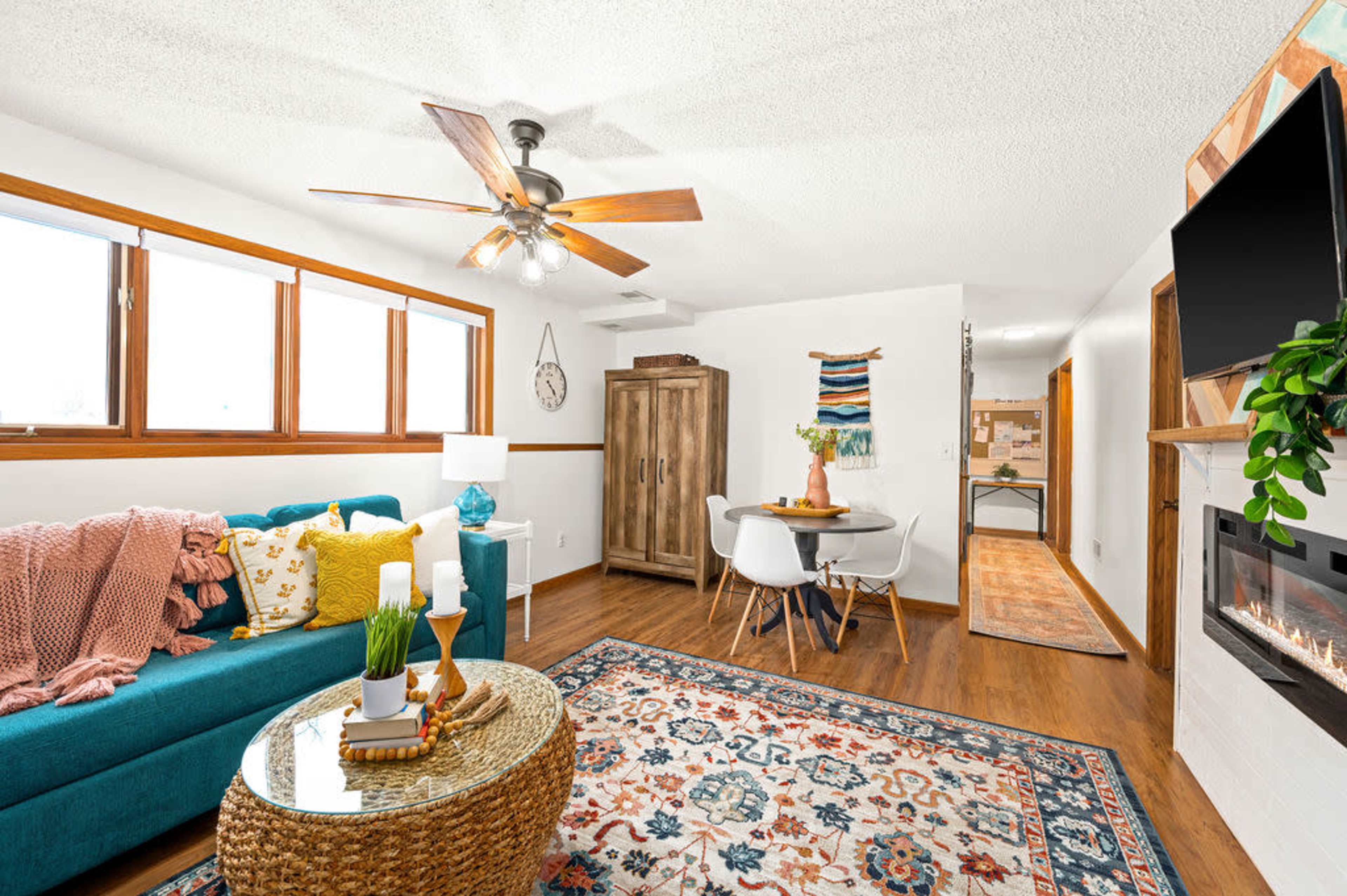 A cozy living room features a blue couch, a round coffee table, and a dining area with a table and chairs, illuminated by natural light from large windows.