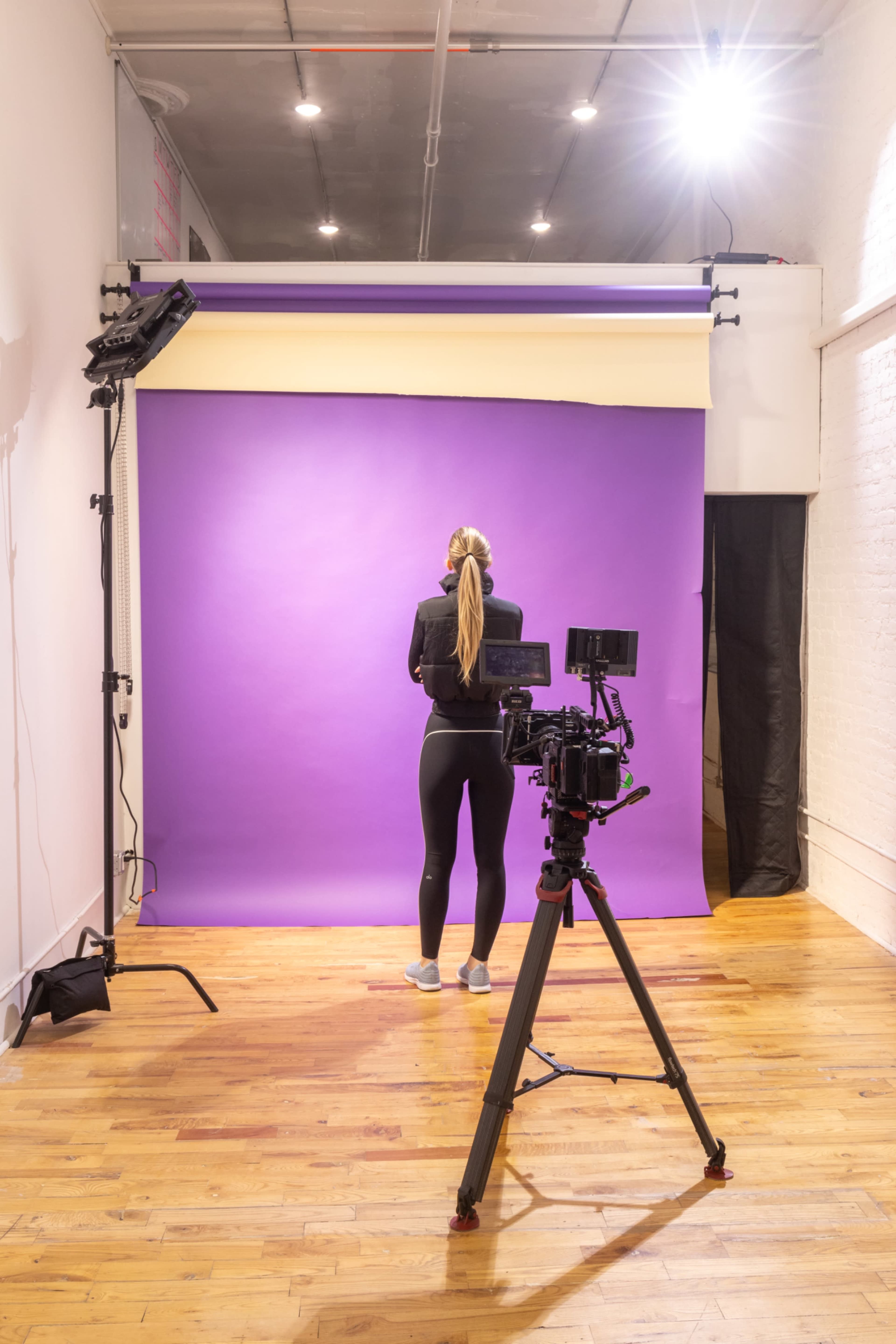 A person stands facing a purple backdrop in a photography studio, with a camera mounted on a tripod nearby.