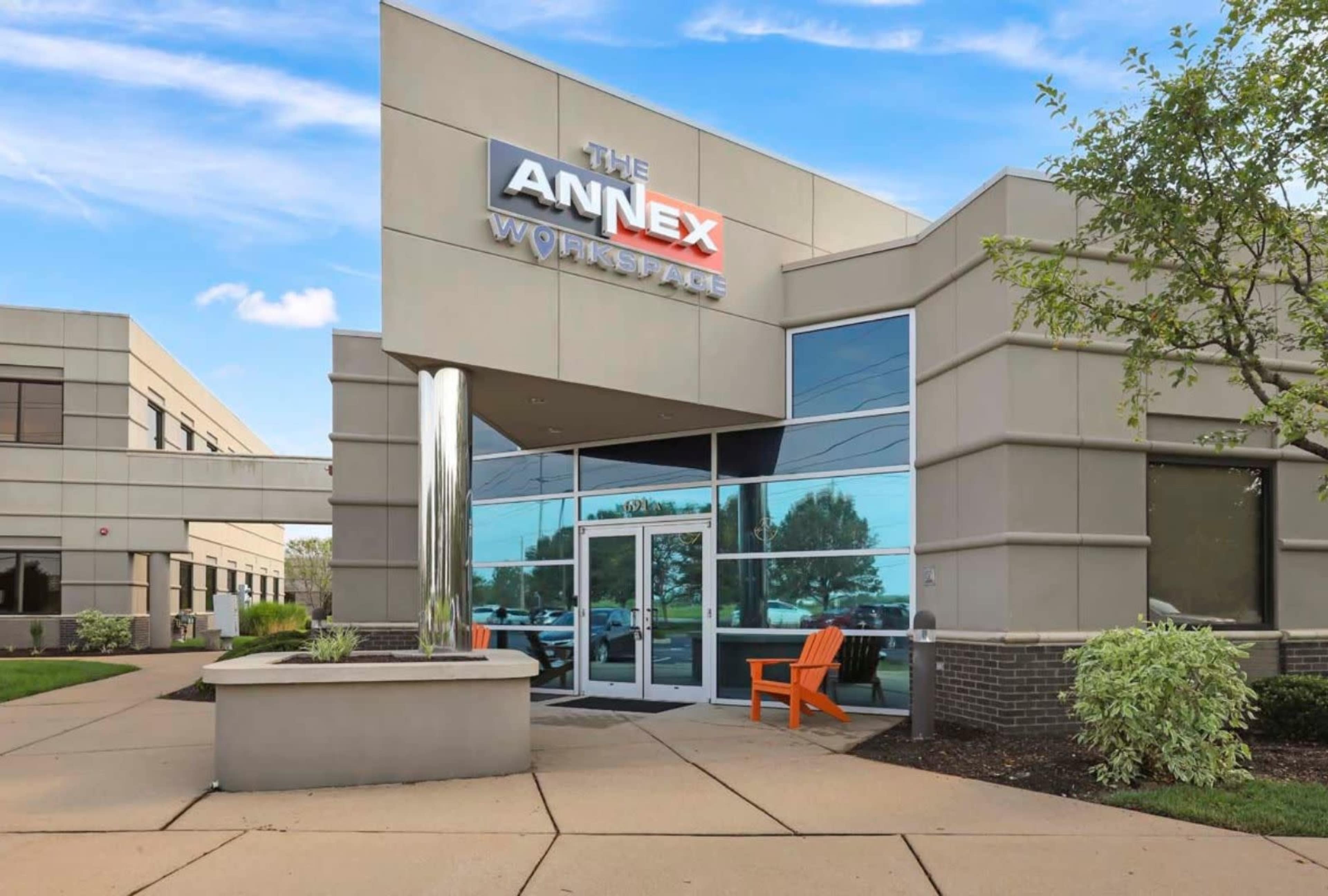 The image shows a modern building entrance with a sign that reads "The Annex Workspace," featuring large windows and an outdoor seating area with an orange chair.