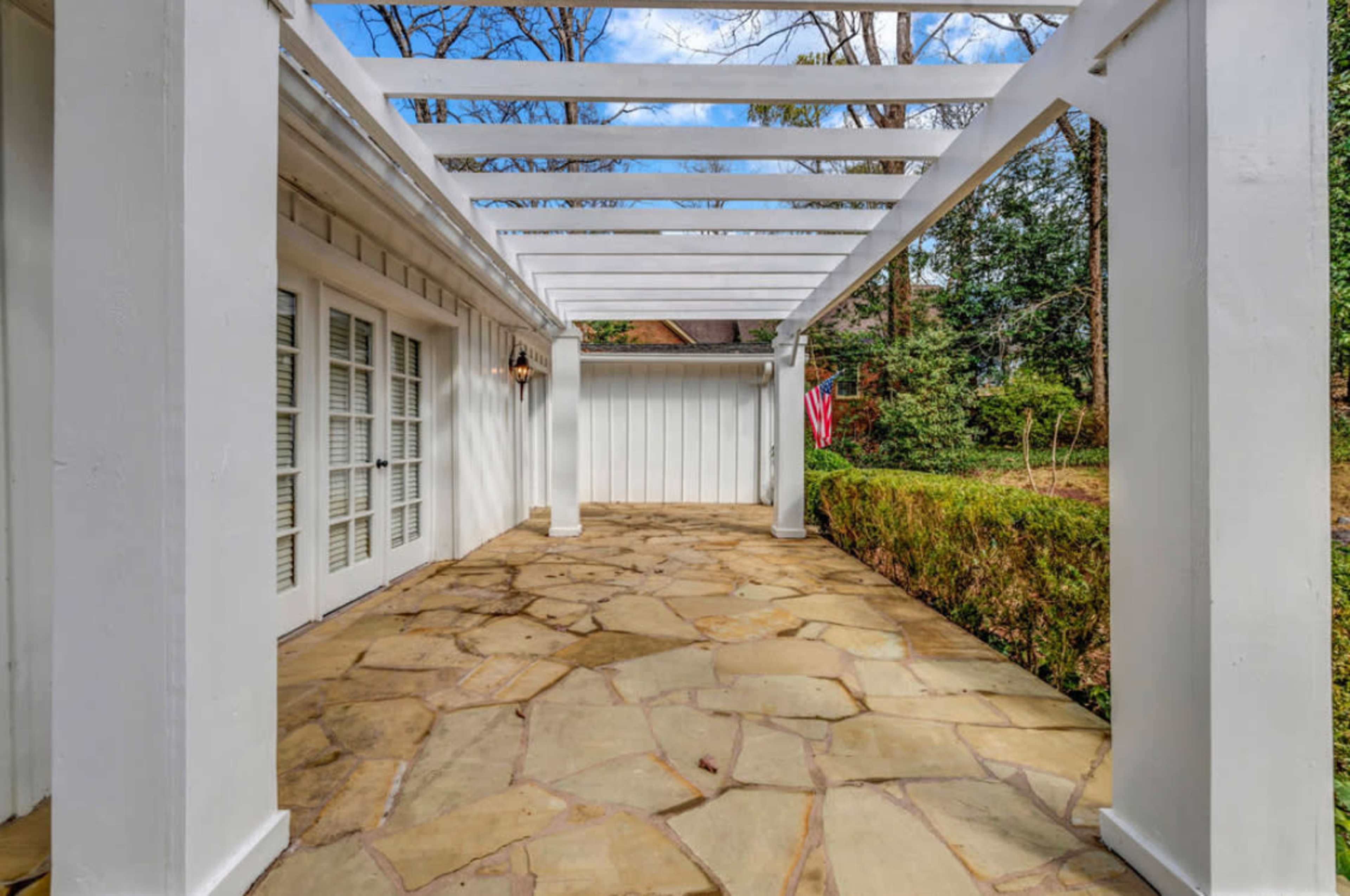 The image shows a stone-paved patio area with a white pergola and greenery in the background.