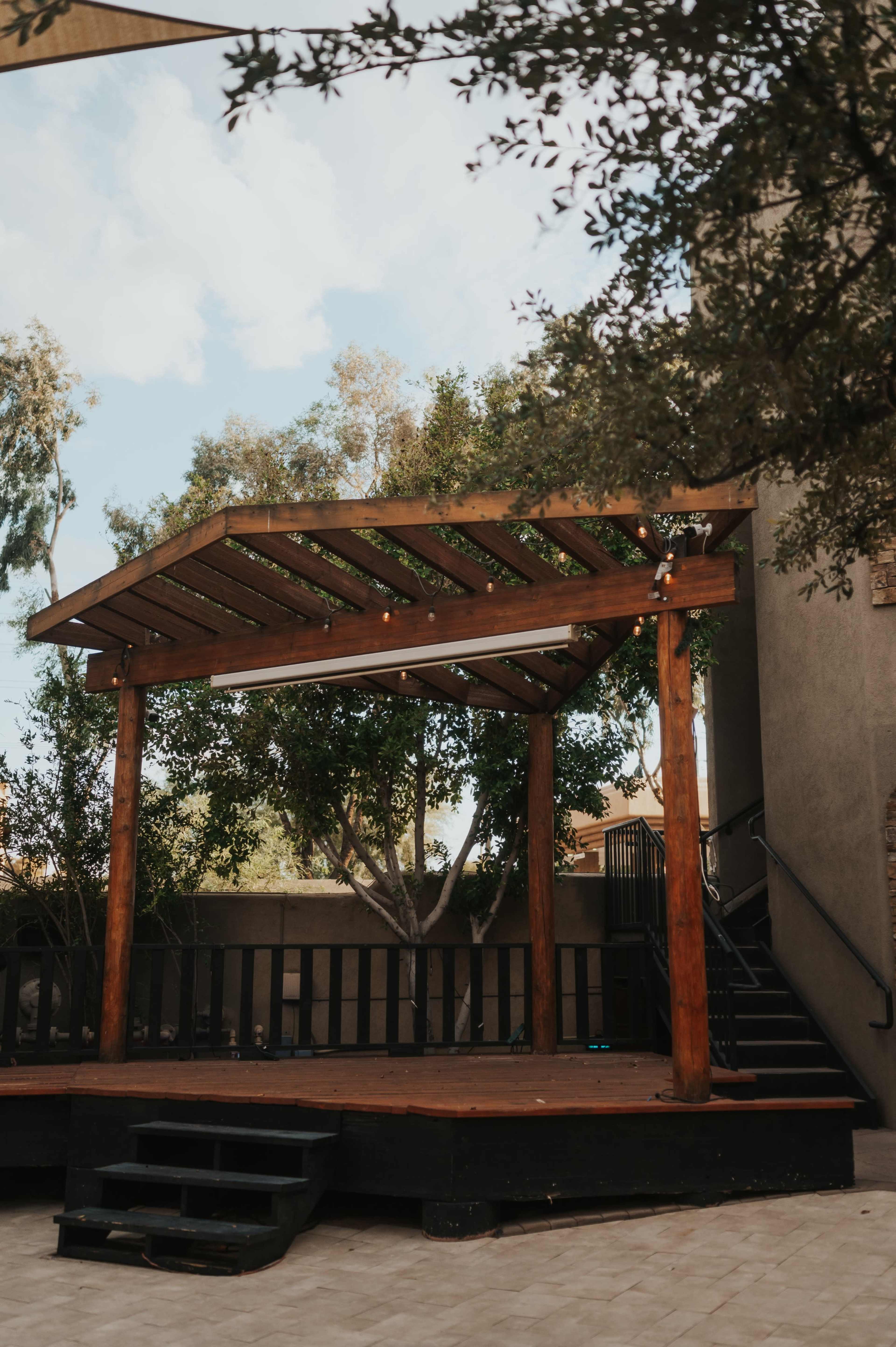 A wooden pergola with string lights is situated in a courtyard surrounded by greenery and a staircase.