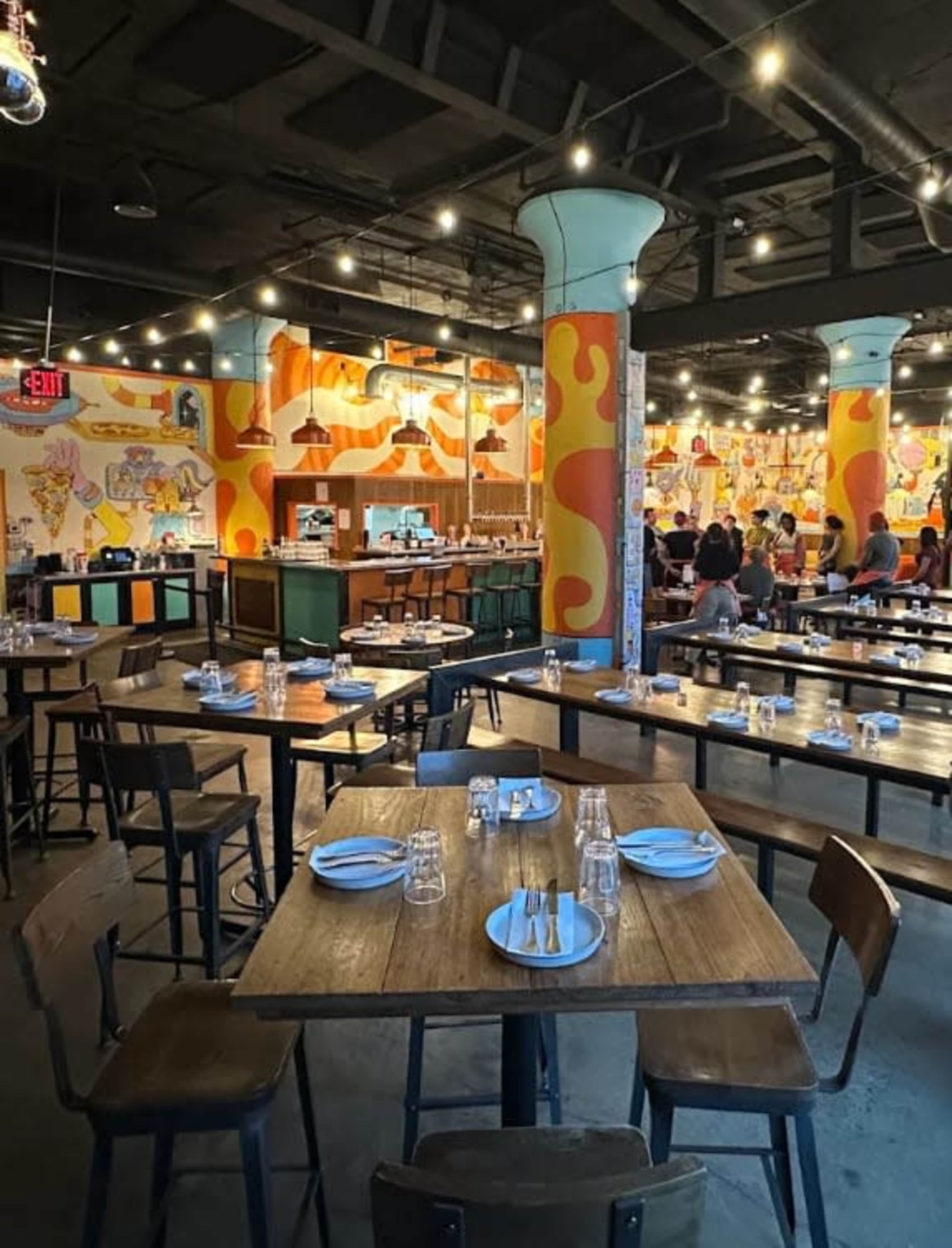 The image shows a vibrant restaurant interior with wooden tables, mismatched chairs, and colorful murals on the walls, illuminated by hanging pendant lights.