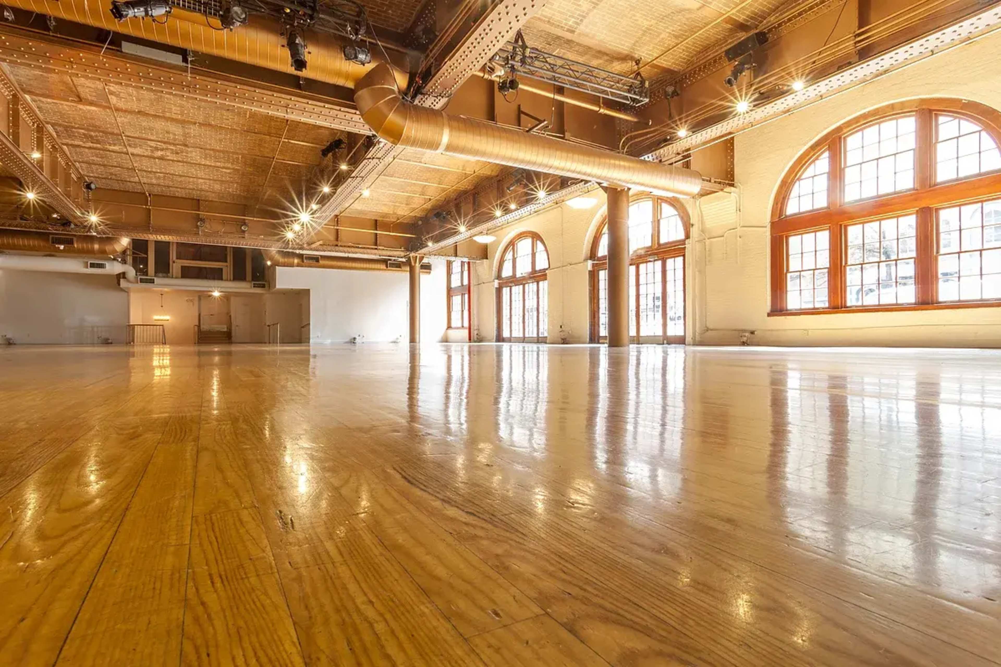 A spacious, empty room with polished wooden floors and large windows allowing natural light.