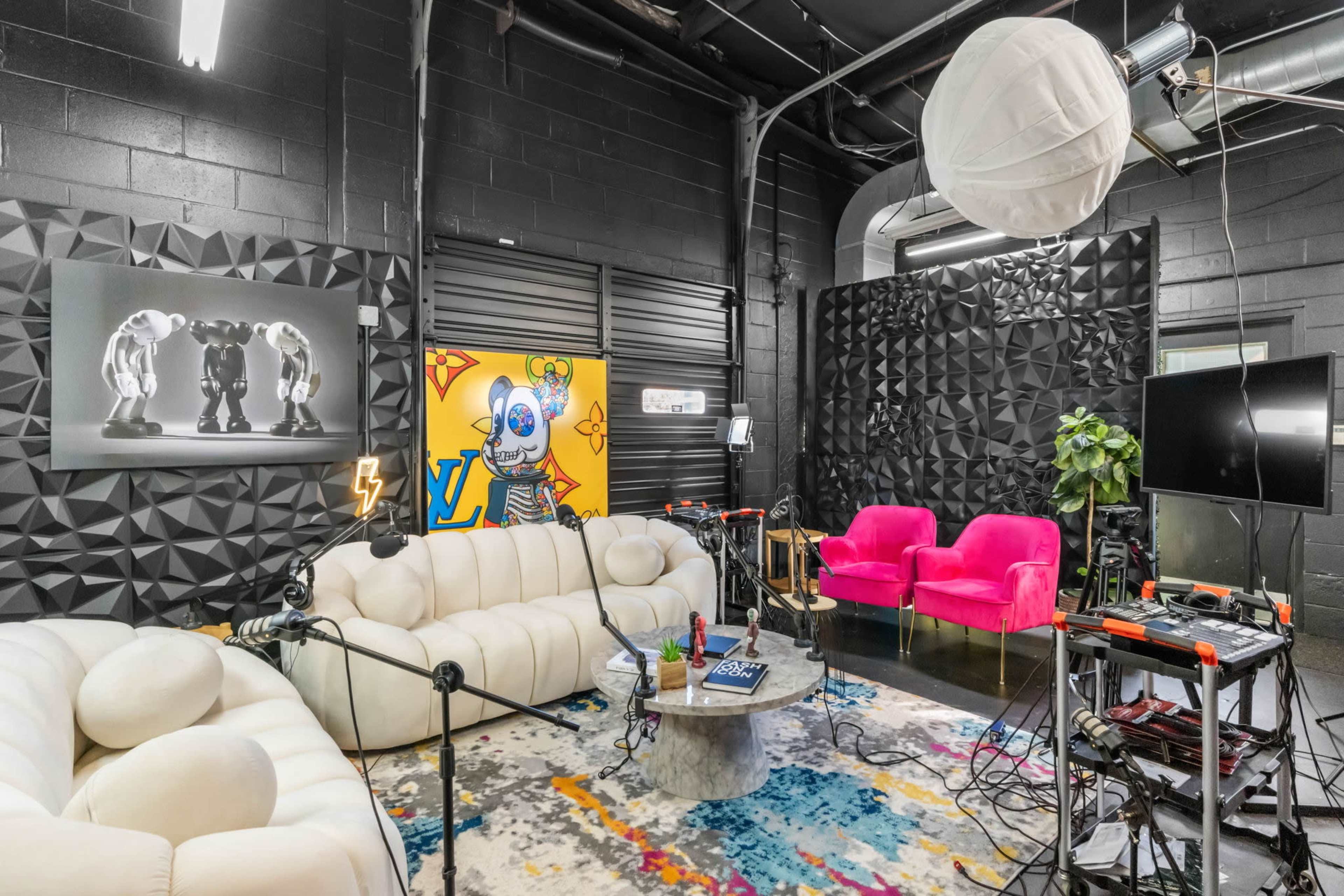 The image shows a modern studio space featuring a light-colored sectional couch, vibrant pink chairs, a multicolored rug, and walls adorned with art and soundproofing panels.