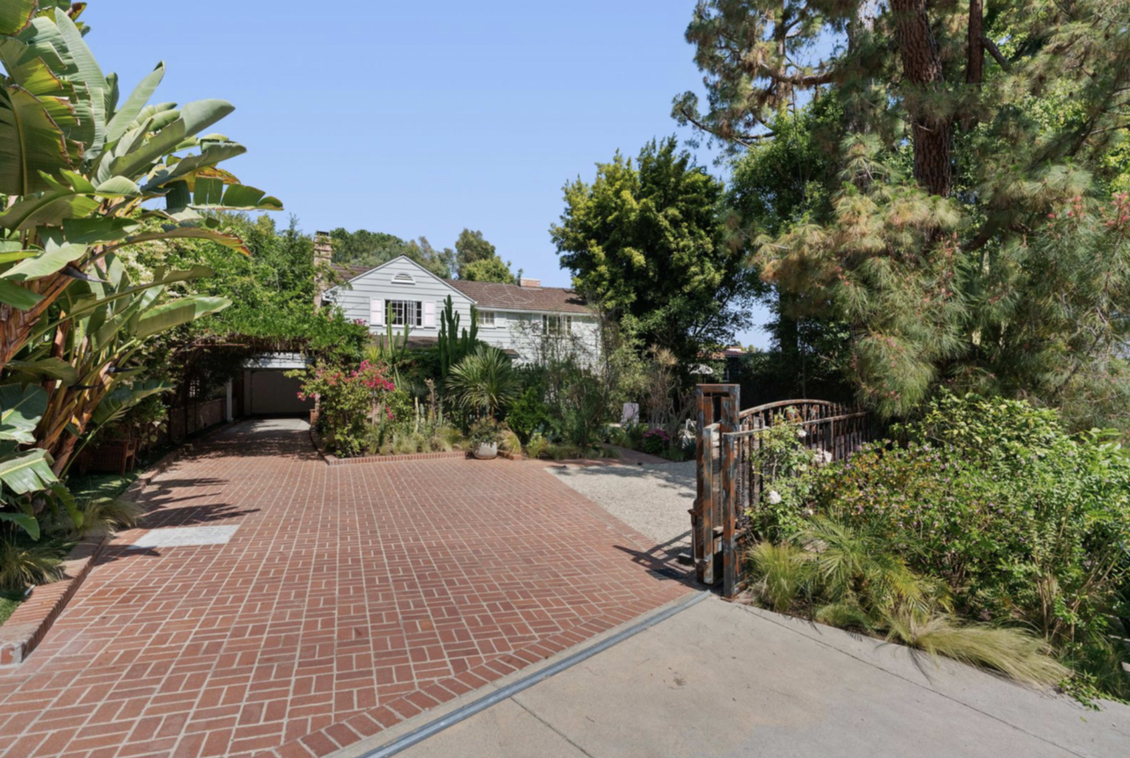 A brick driveway leads to a house surrounded by lush greenery and various plants.