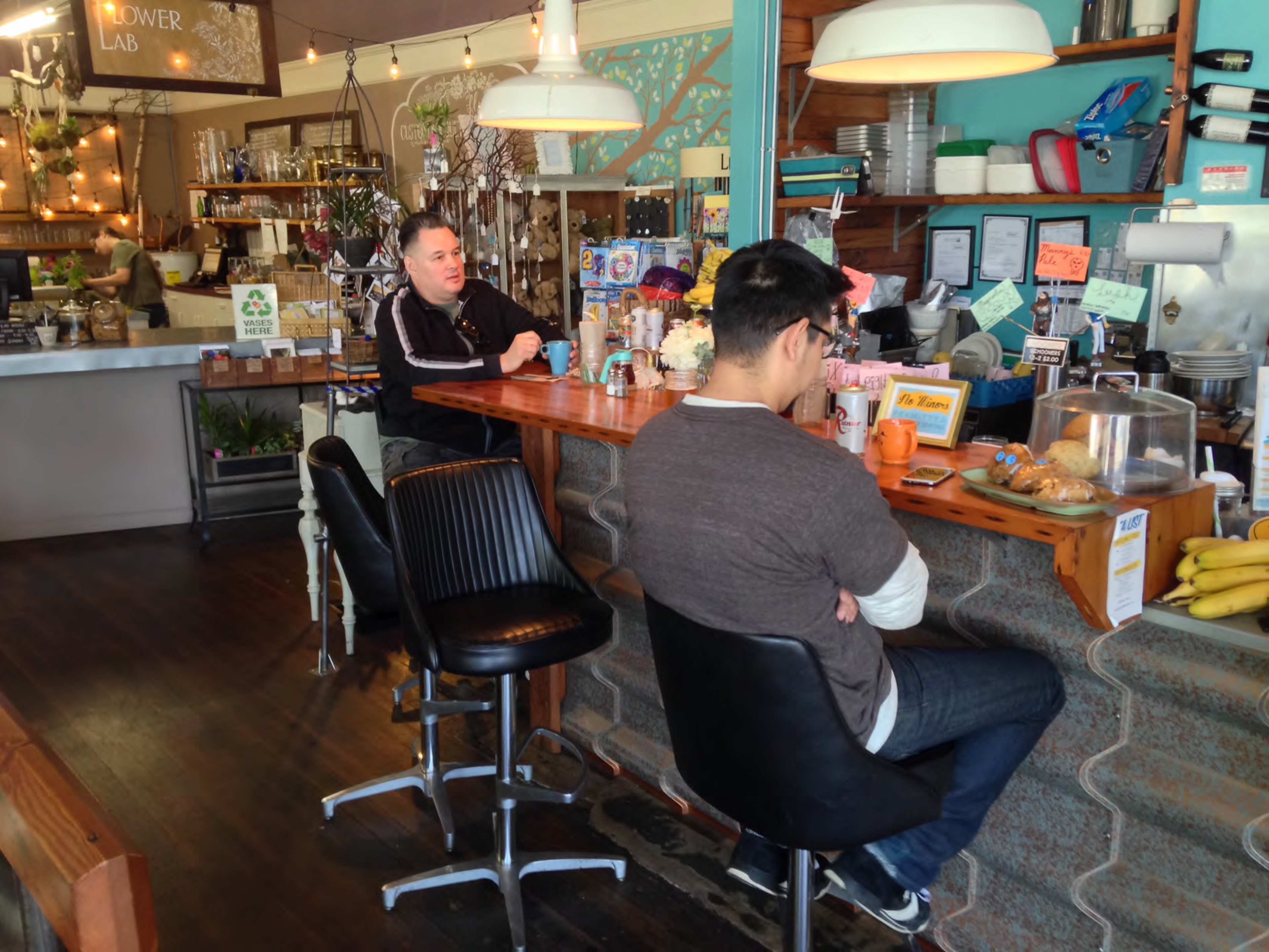 A barista stands behind a counter in a café with two customers seated on stools, surrounded by decor and various items on display.