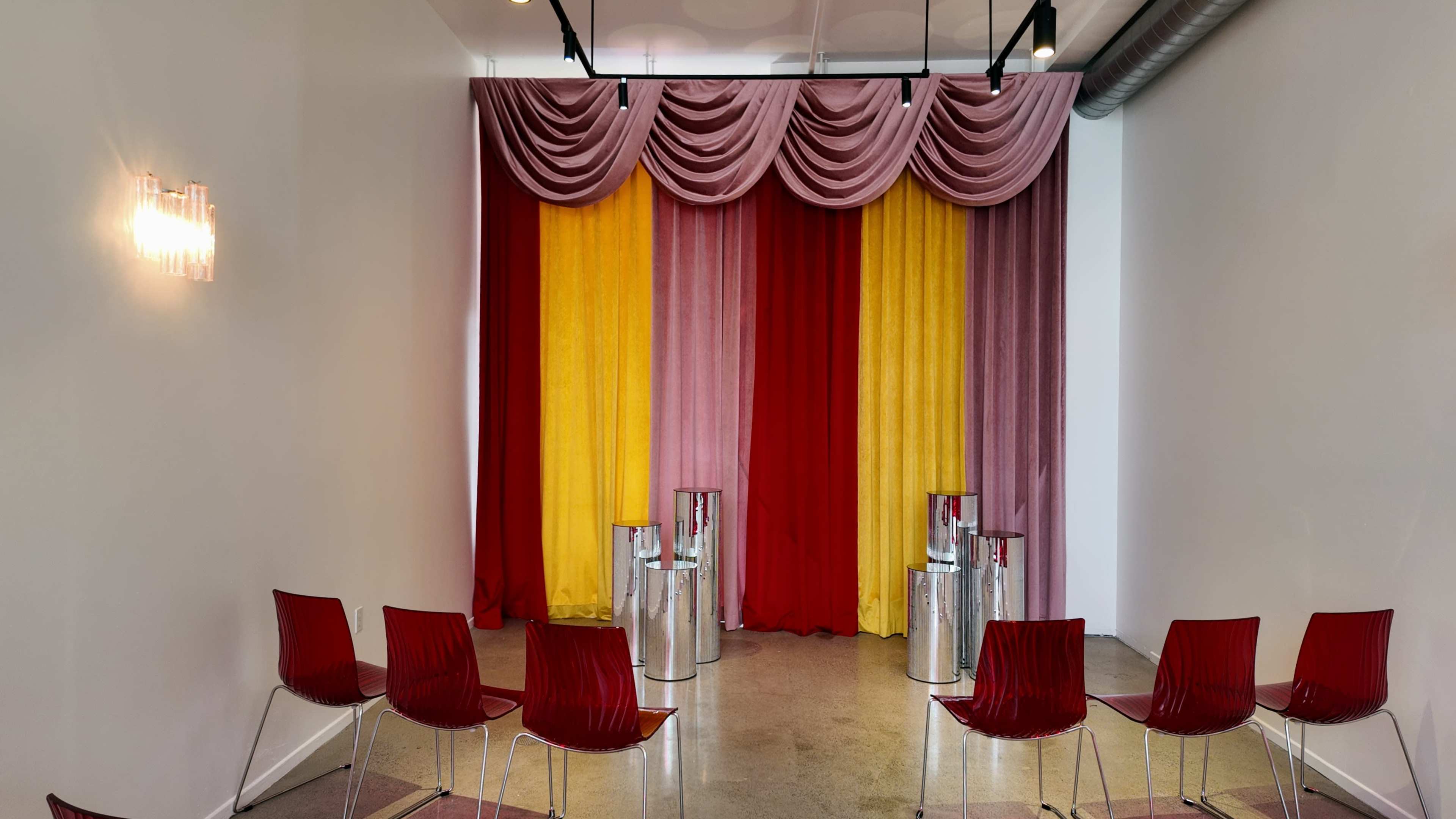 A room with a colorful curtain backdrop in pink, yellow, and red, with several red chairs arranged in a semicircle in front of it.