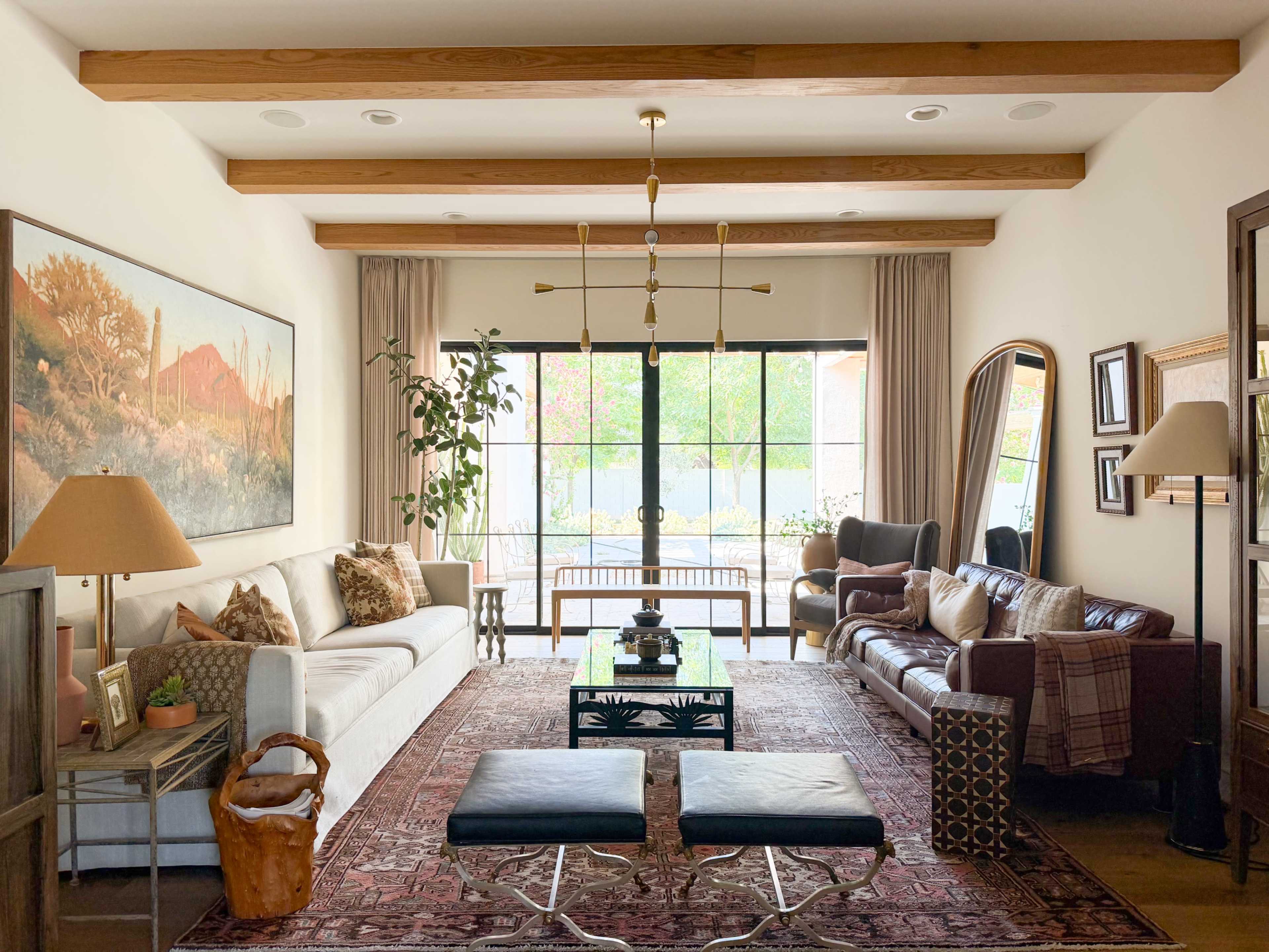 The living room features a white sofa and a brown leather couch, complemented by a large window, decorative lamps, and a combination of artwork and plants.
