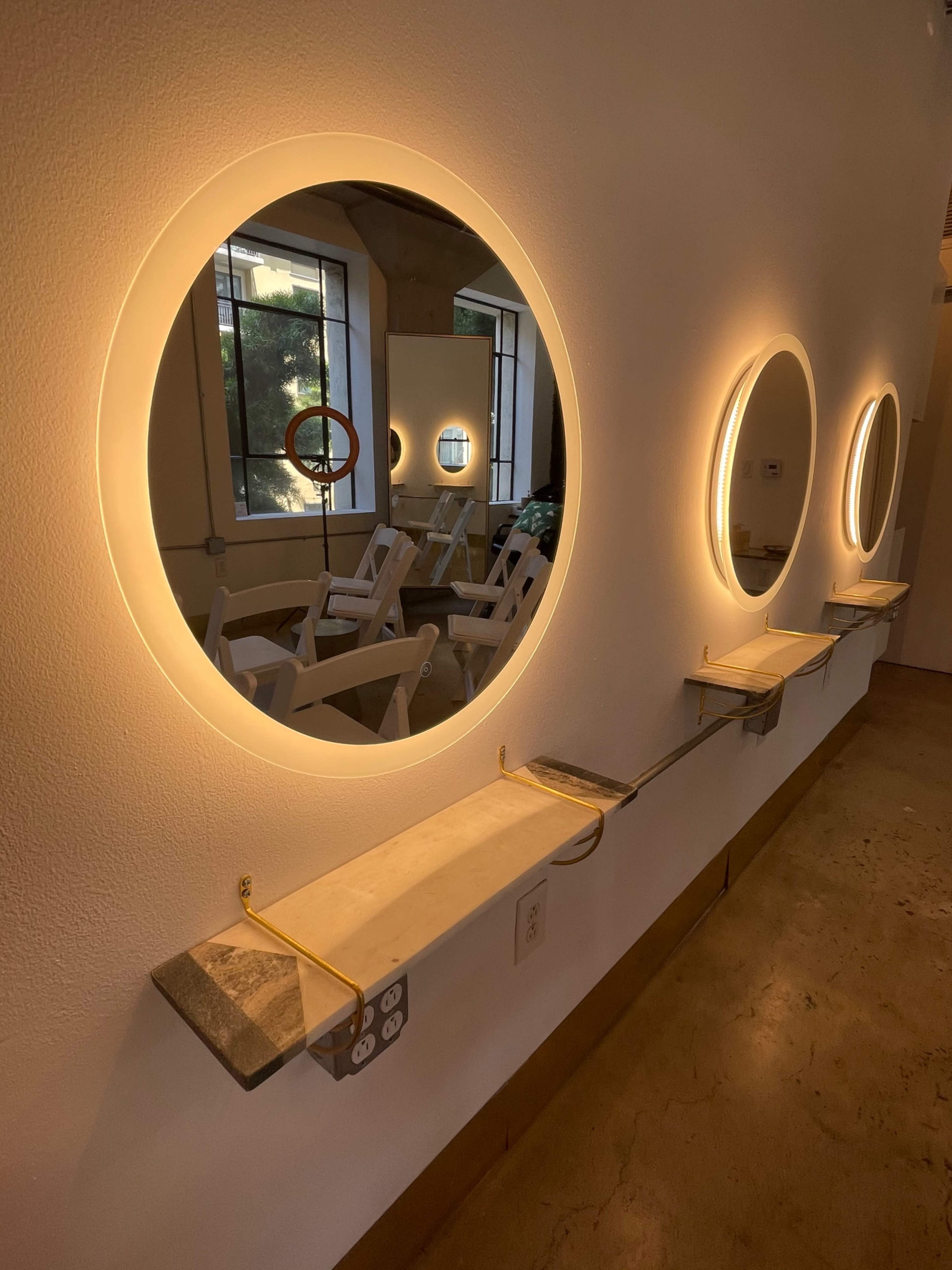 The image shows a row of circular mirrors with integrated lighting mounted on a wall, accompanied by marble shelves beneath each mirror.