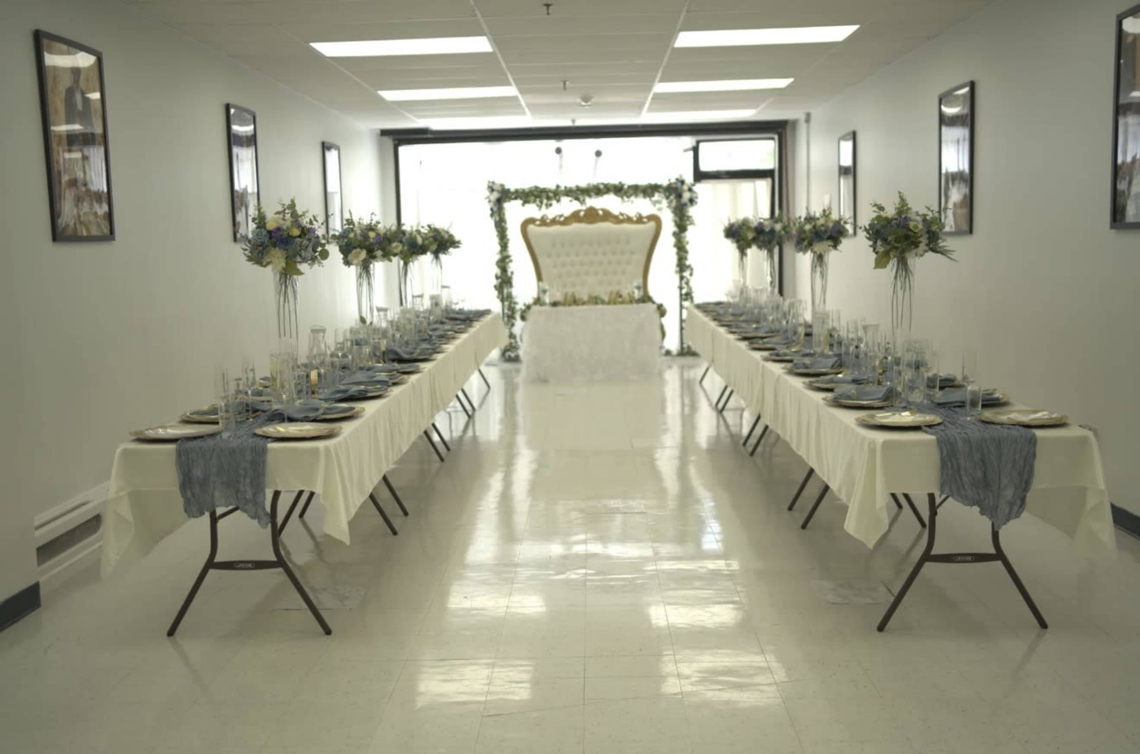 A long banquet hall is set up with two parallel tables adorned with white tablecloths, gold plates, and floral centerpieces, leading to a decorated backdrop.