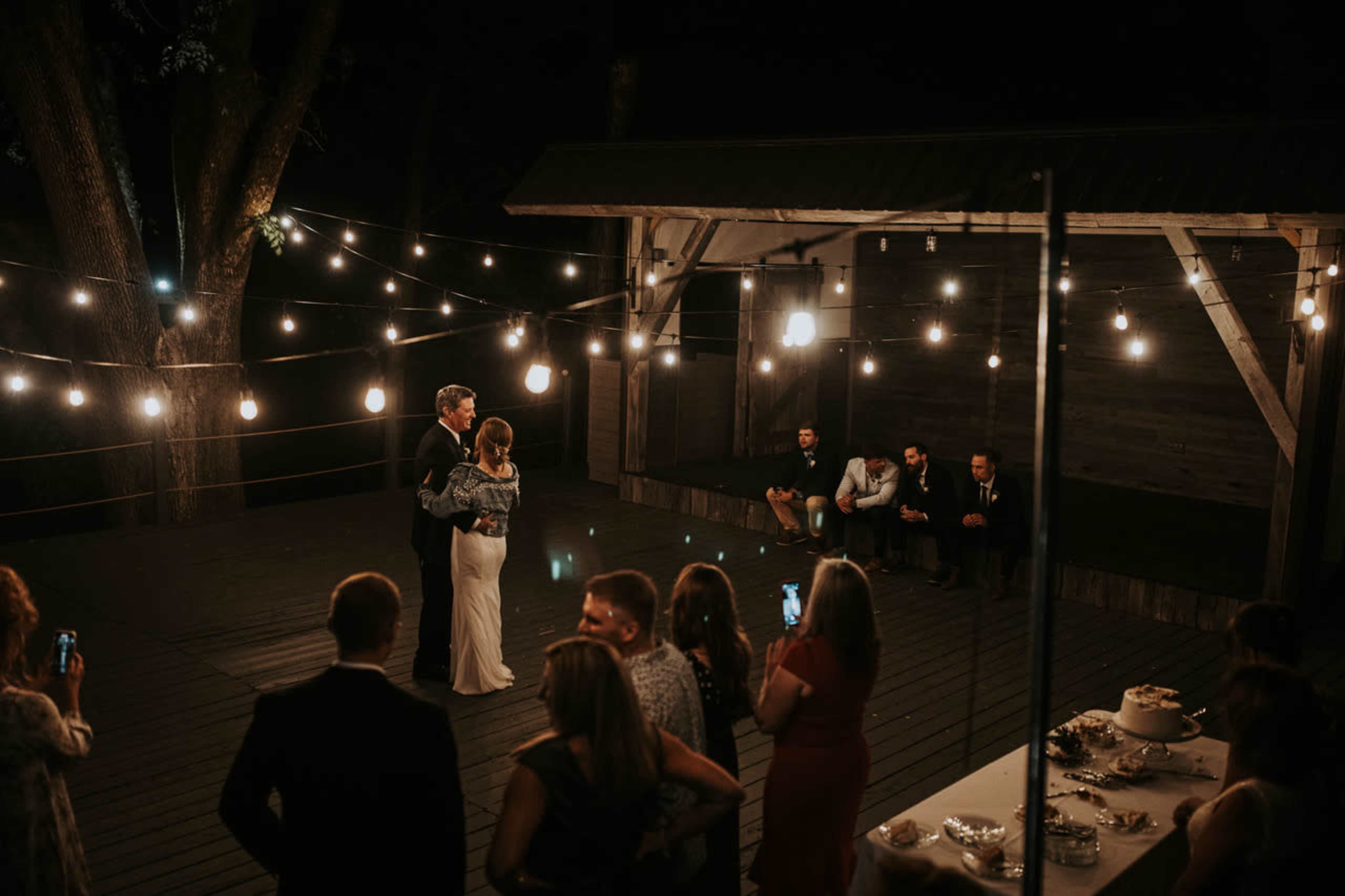 A couple dances under string lights on a wooden deck while guests watch and take photos.