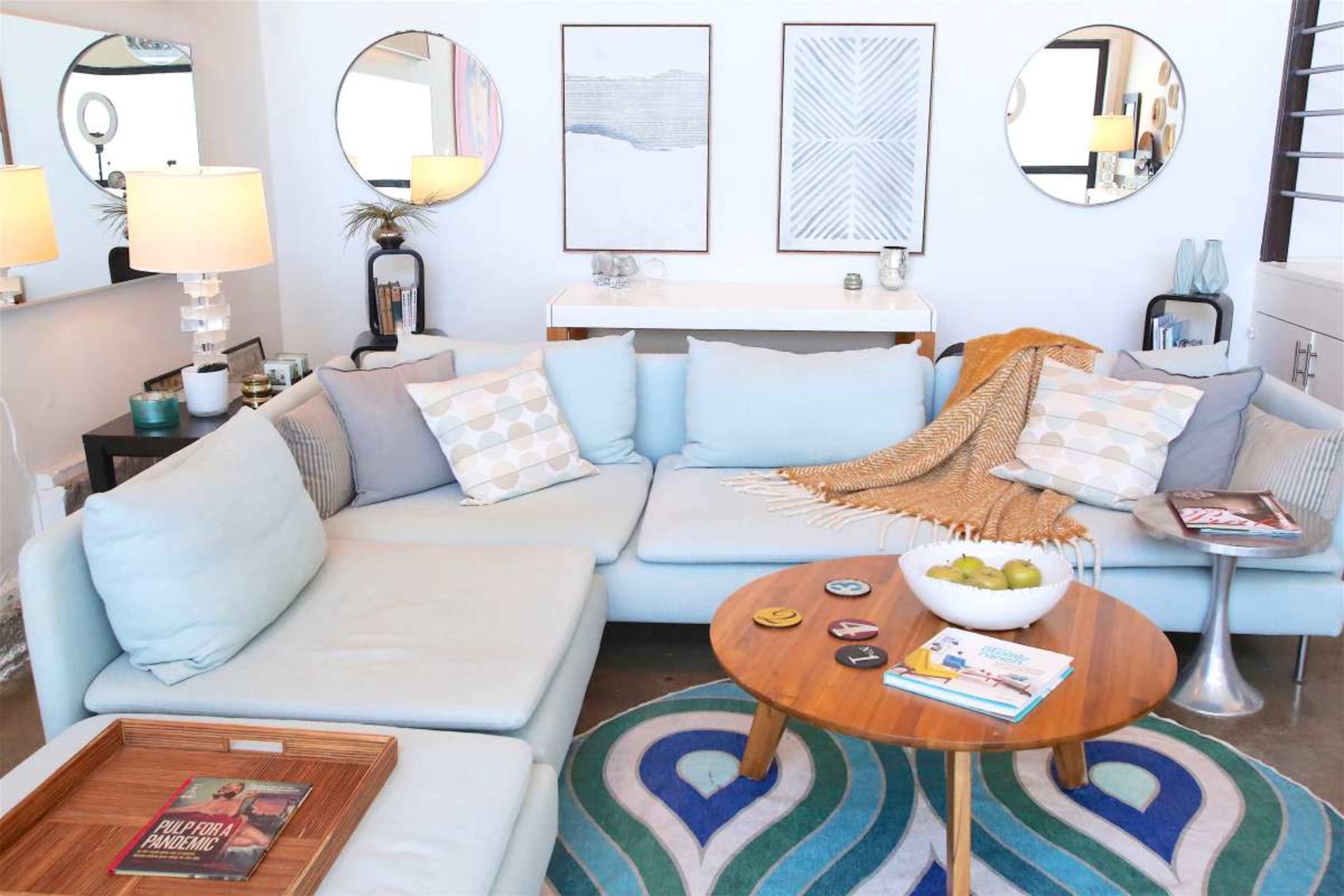 The image shows a modern living room with a light blue sectional sofa, decorative pillows, a round wooden coffee table, and circular mirrors on the wall.