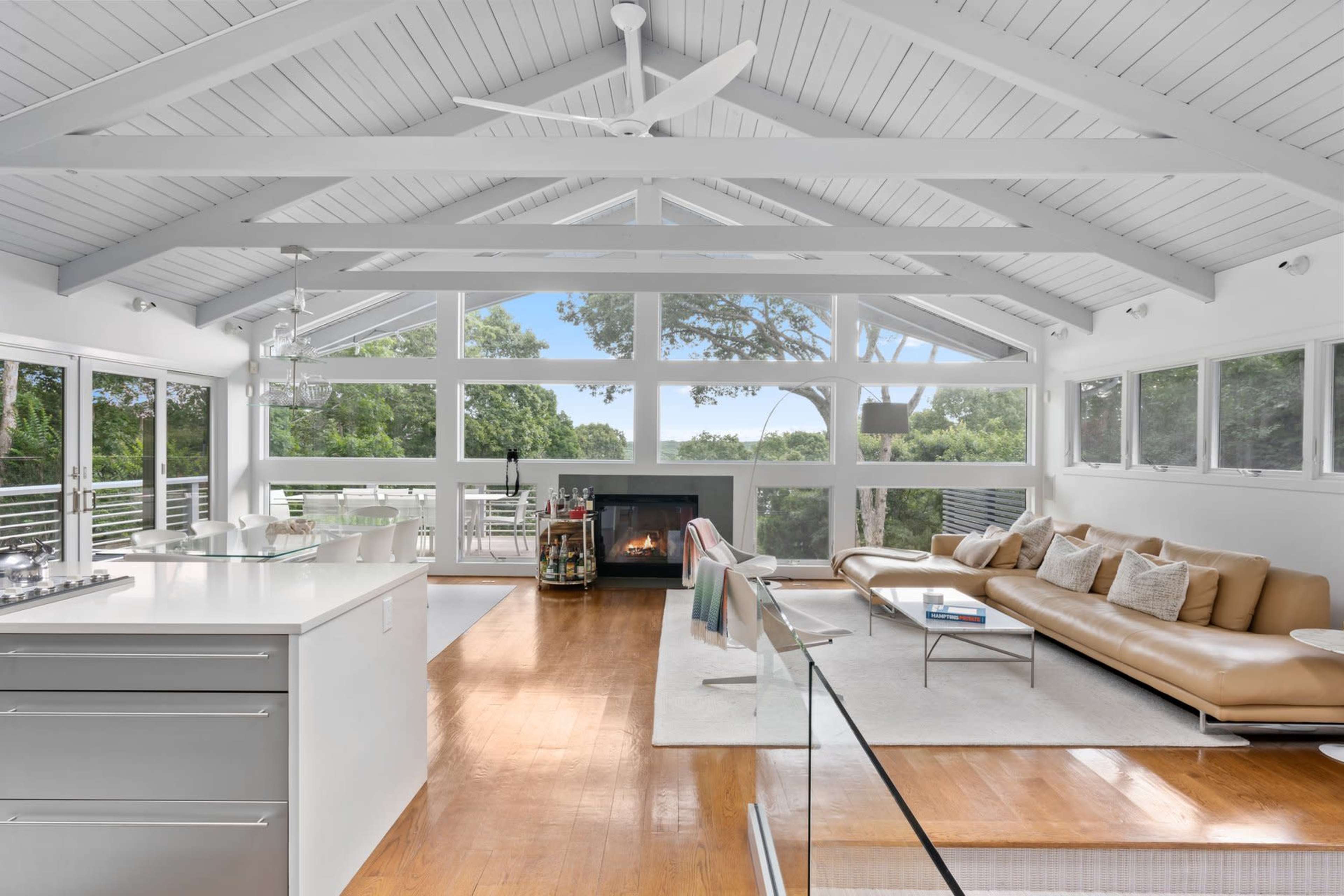 A modern living room with large windows, a fireplace, and a view of greenery outside.