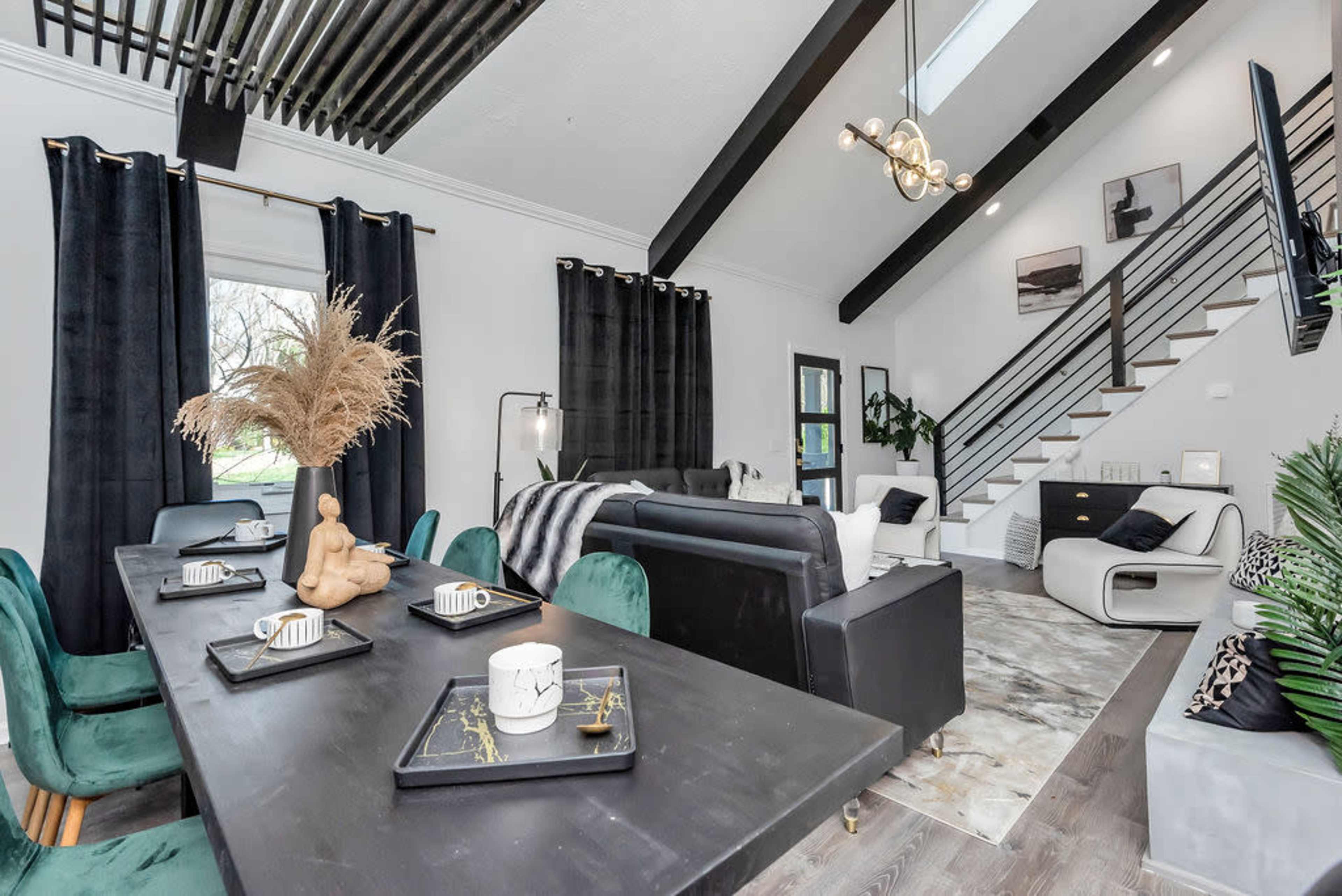 A modern living space features a dining area with a dark table and green chairs, a black sofa, and a staircase leading to an upper level, adorned with plants and decorative accents.