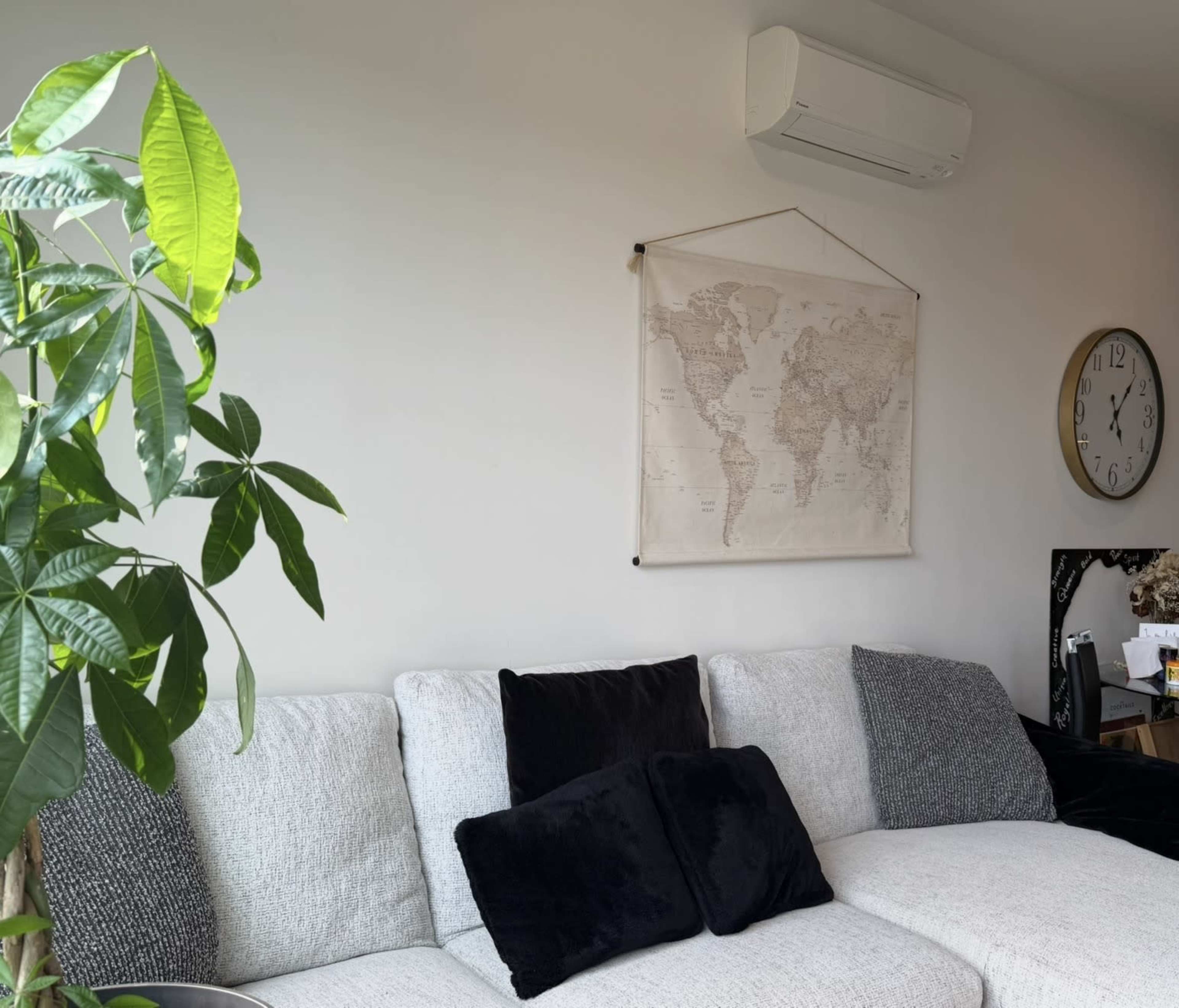 The image shows a cozy living room corner with a sectional sofa, a large wall map hanging above it, a plant on the left, and a clock on the wall.