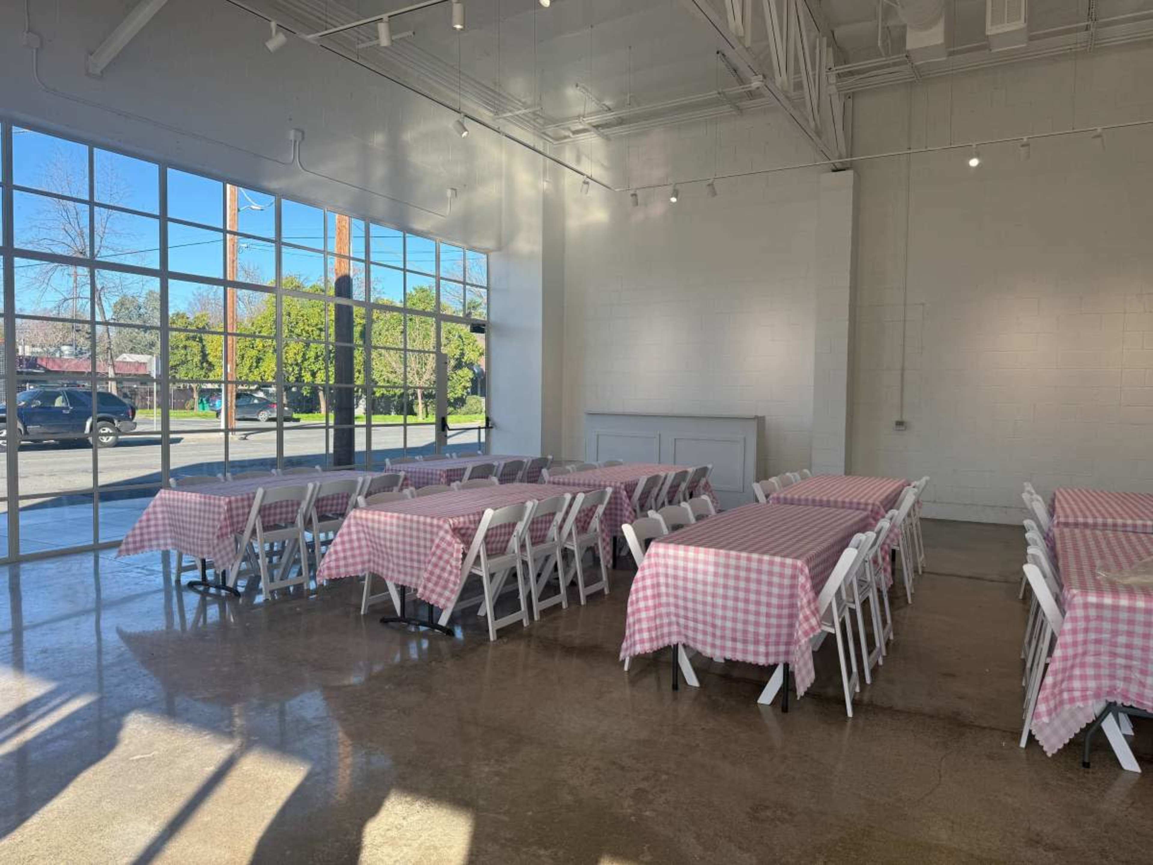 The image shows a spacious indoor venue with long tables covered in pink-and-white checkered tablecloths set up near large windows letting in natural light.