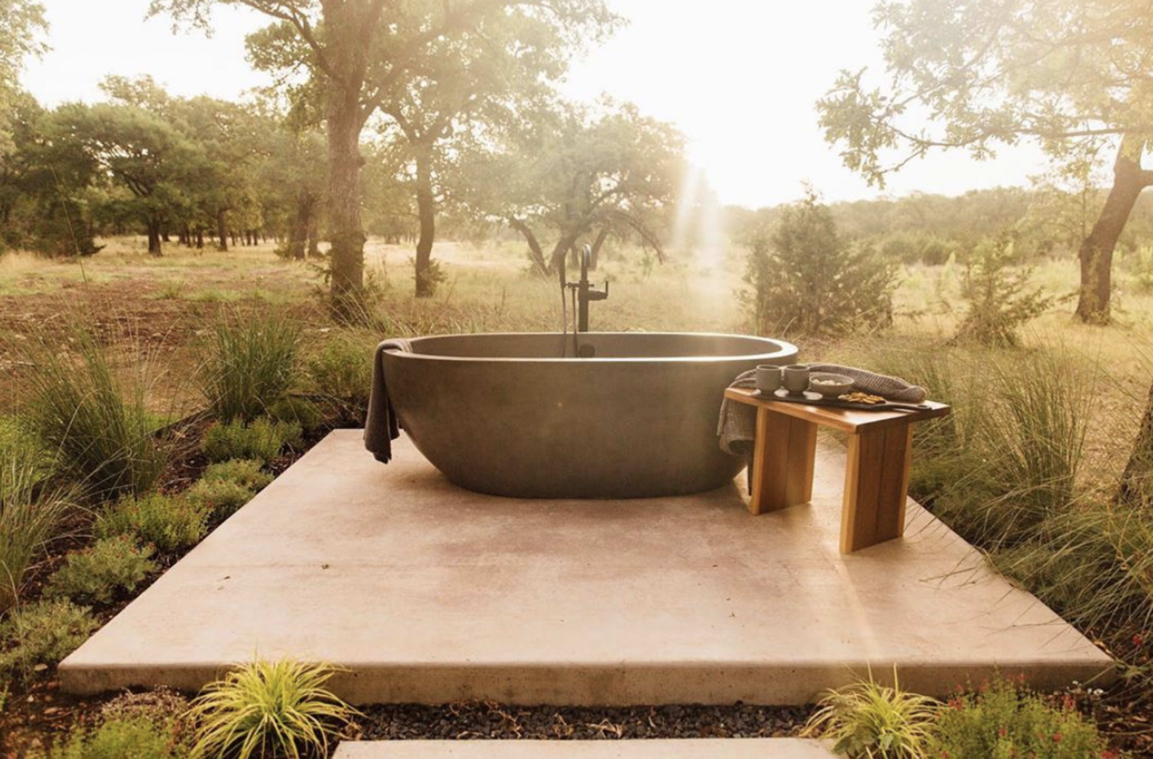 A modern freestanding bathtub sits on a concrete platform surrounded by greenery in a natural setting.