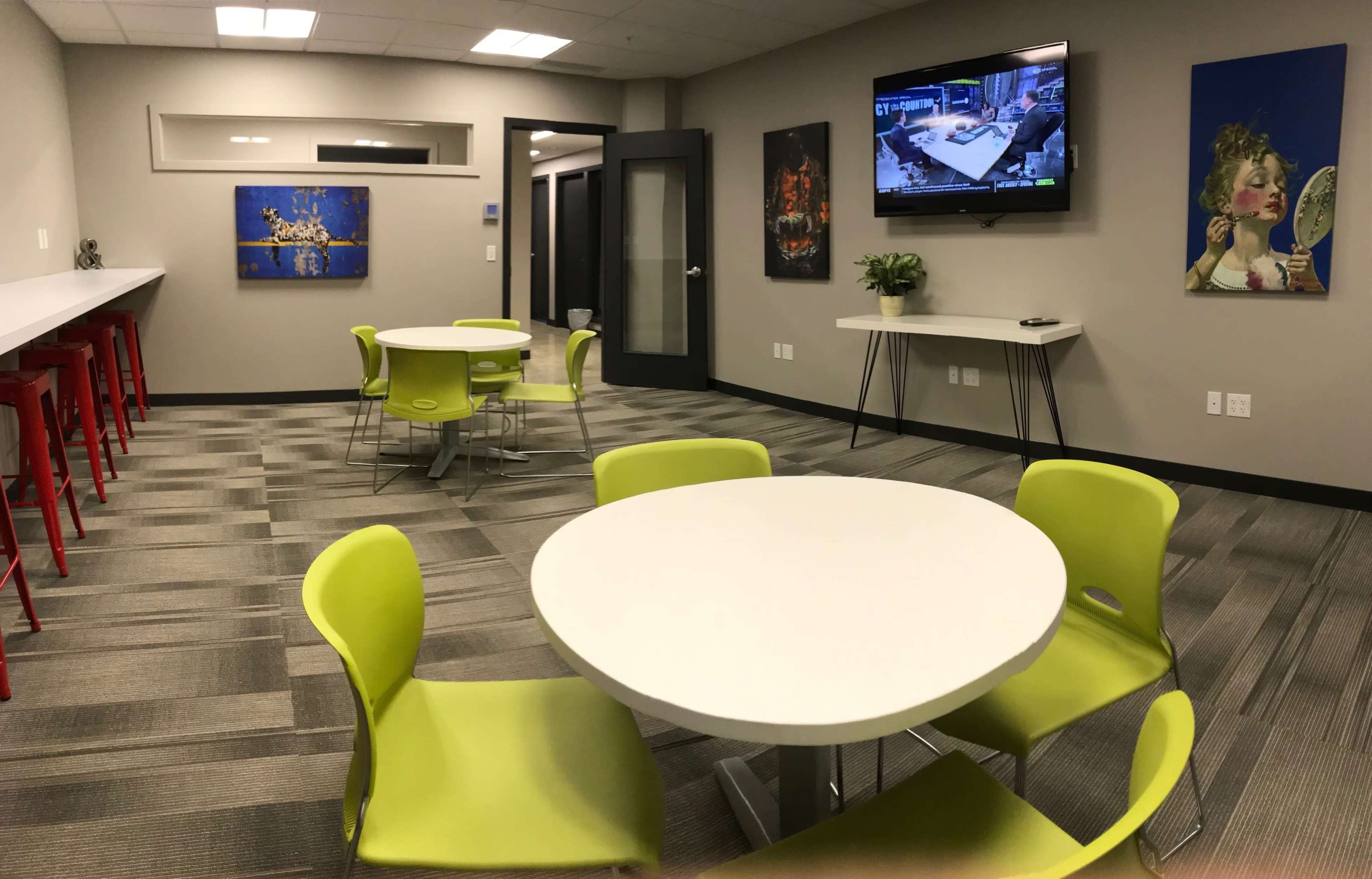 A modern office space featuring green chairs around circular white tables, a television mounted on the wall, and colorful artwork displayed.