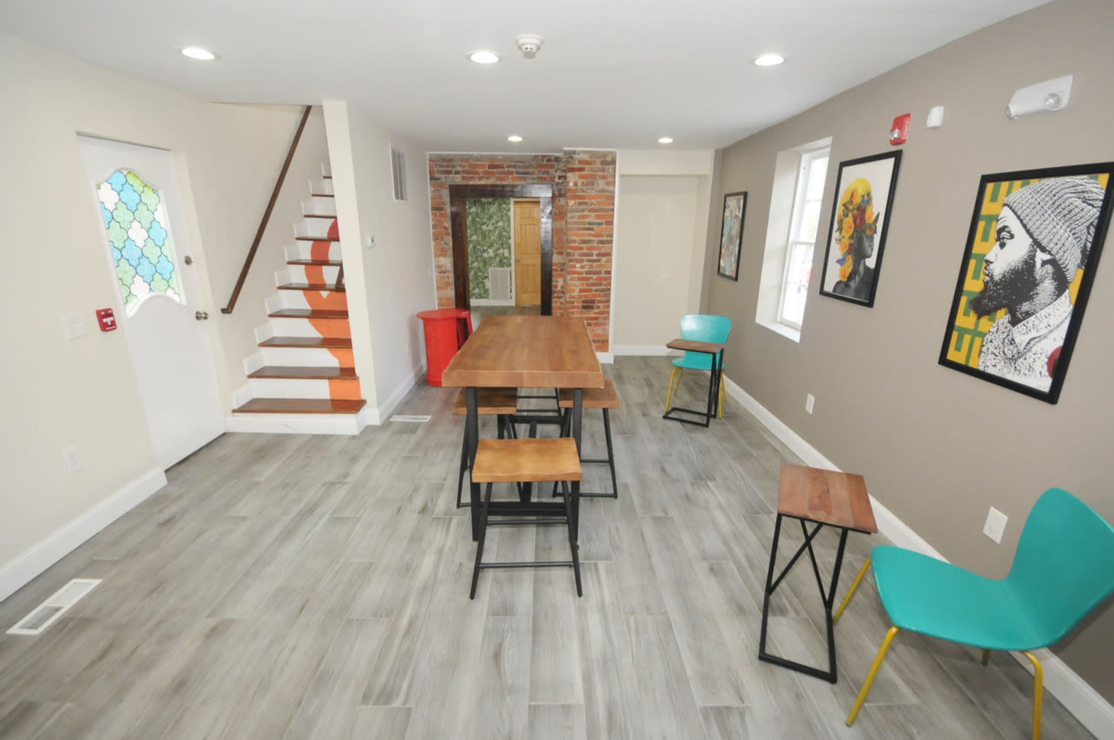 A modern interior space with a wooden dining table and chairs, a staircase leading to an upper level, and exposed brick on one wall.