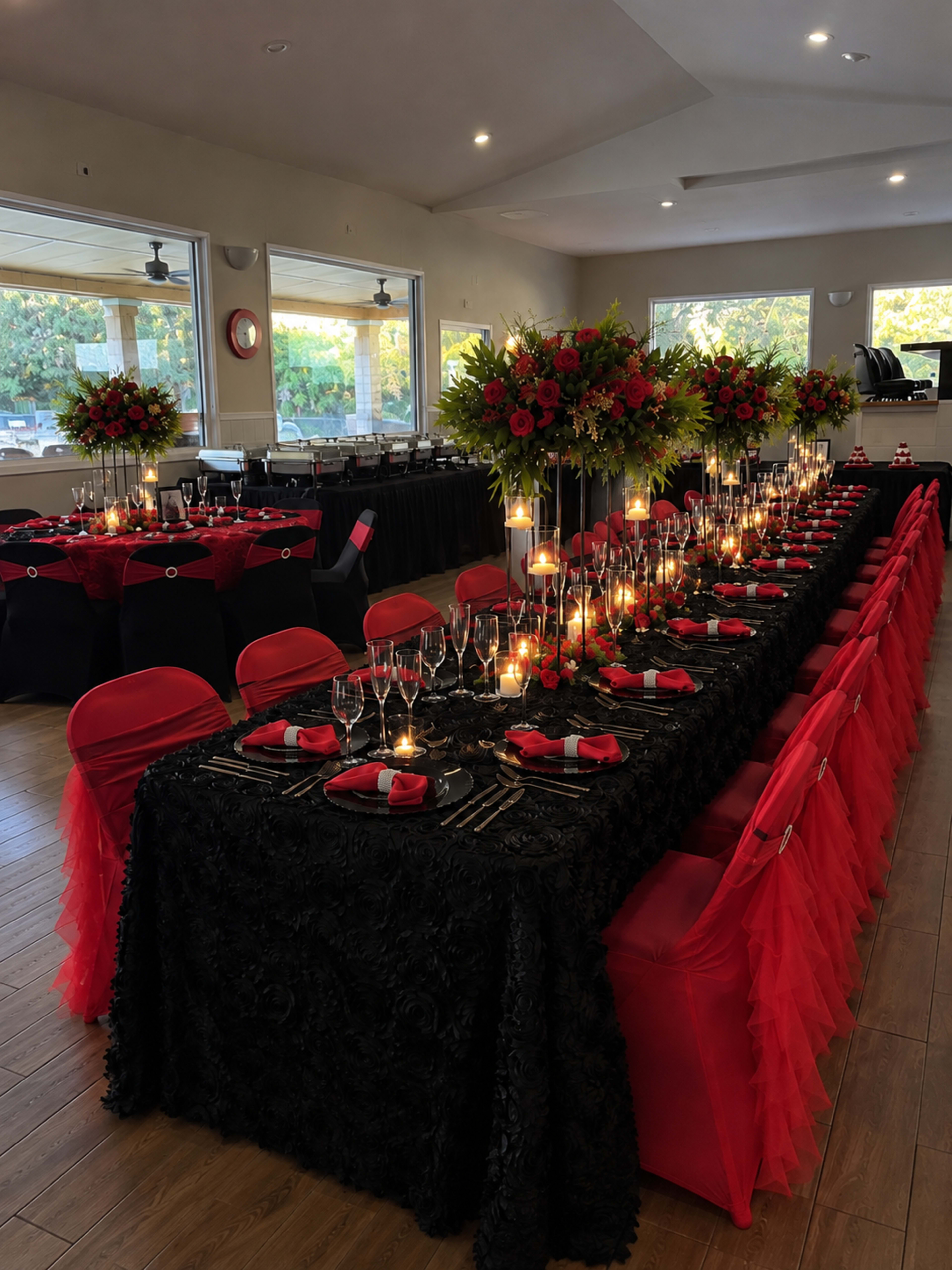 The image shows a banquet hall set for an event, featuring a long table adorned with black and red decorations, centerpieces of flowers, and candles, surrounded by elegantly draped chairs.