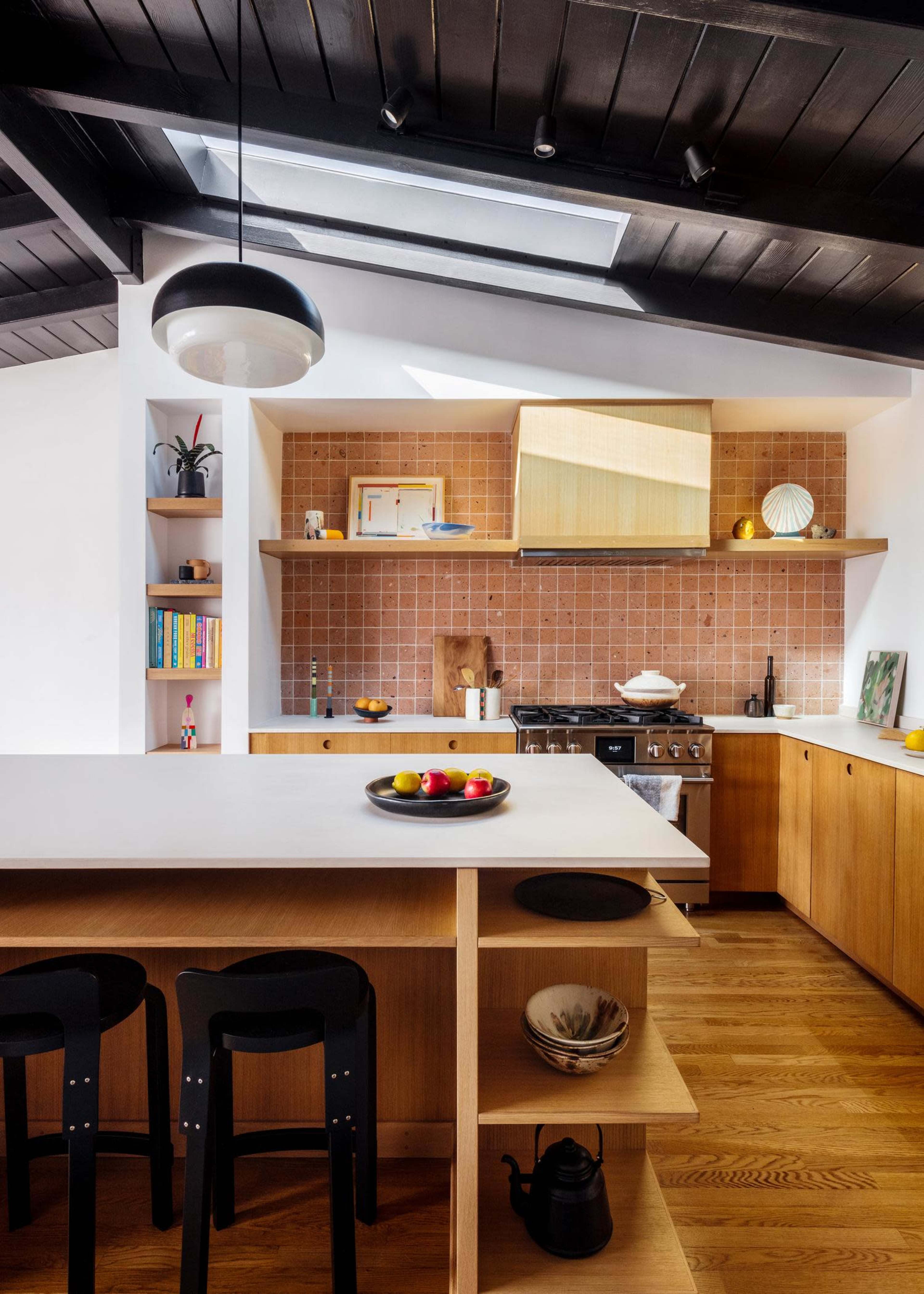 The image shows a modern kitchen with a central island, wooden cabinets, a skylight, and a mix of decorative items and fruits on display.