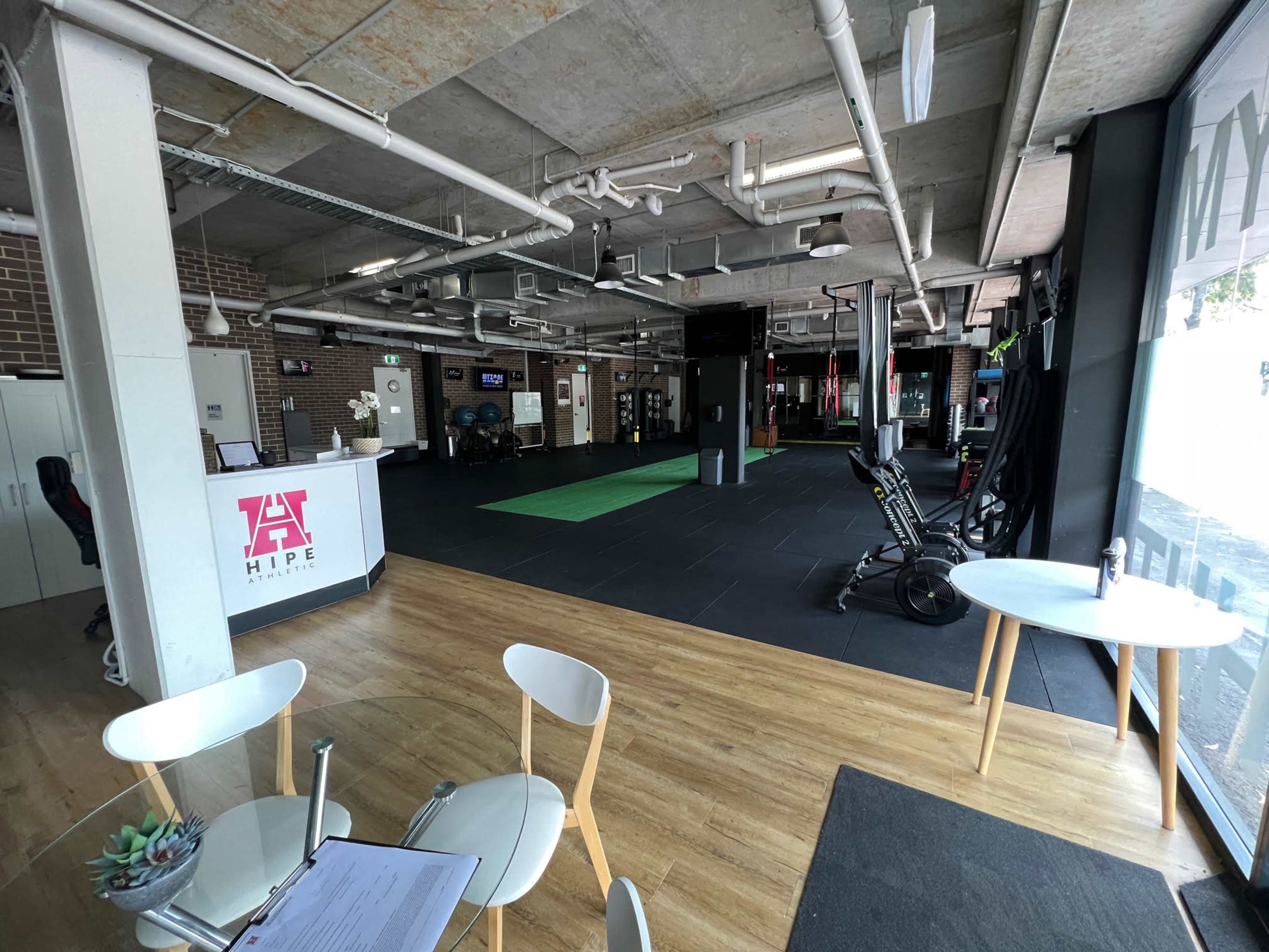 A modern fitness facility featuring an open gym area with exercise equipment, a reception desk, and wooden flooring.