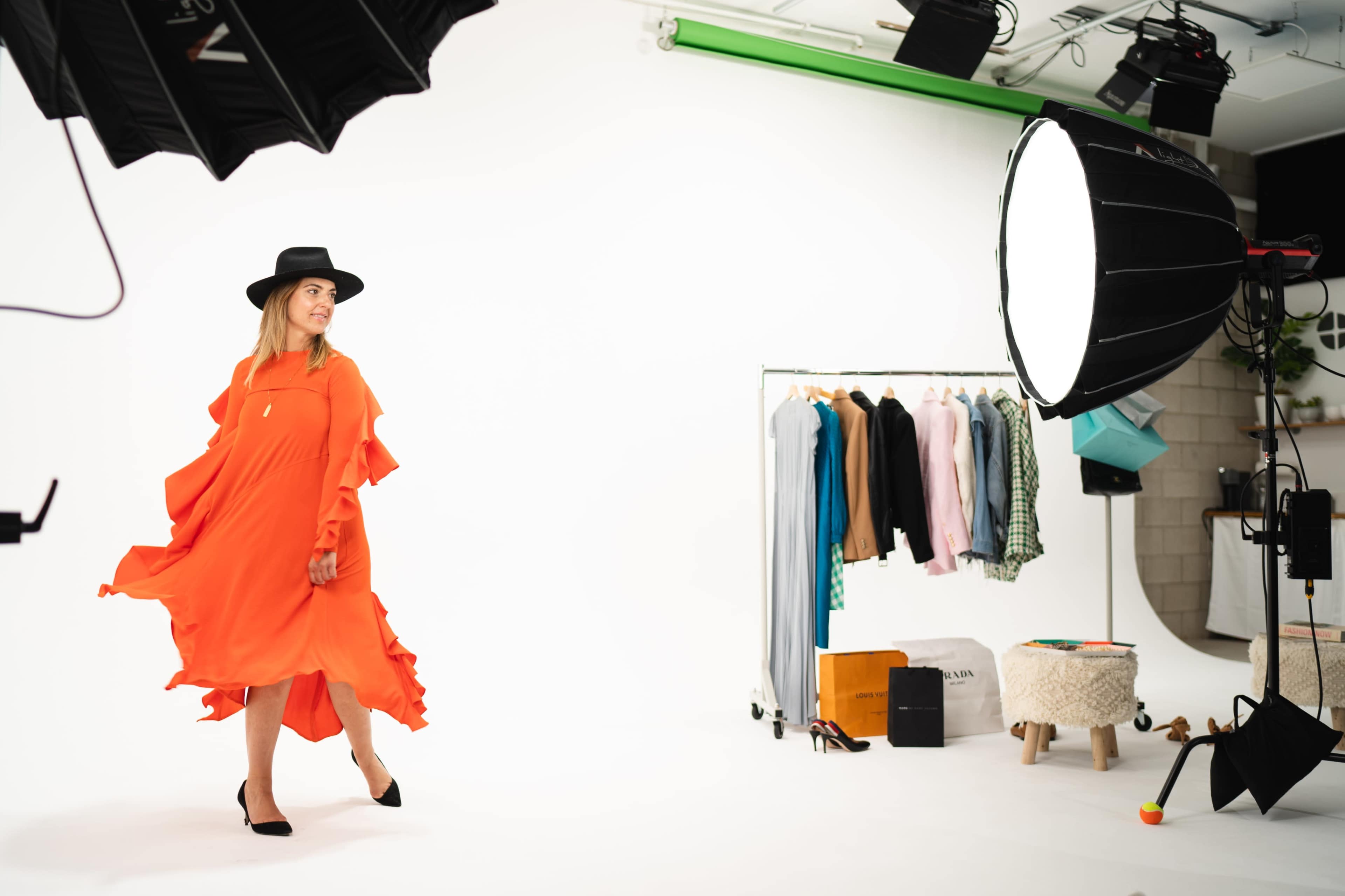A model wearing an orange dress and a black hat walks on a white backdrop, with a clothing rack and photography lights in the background.