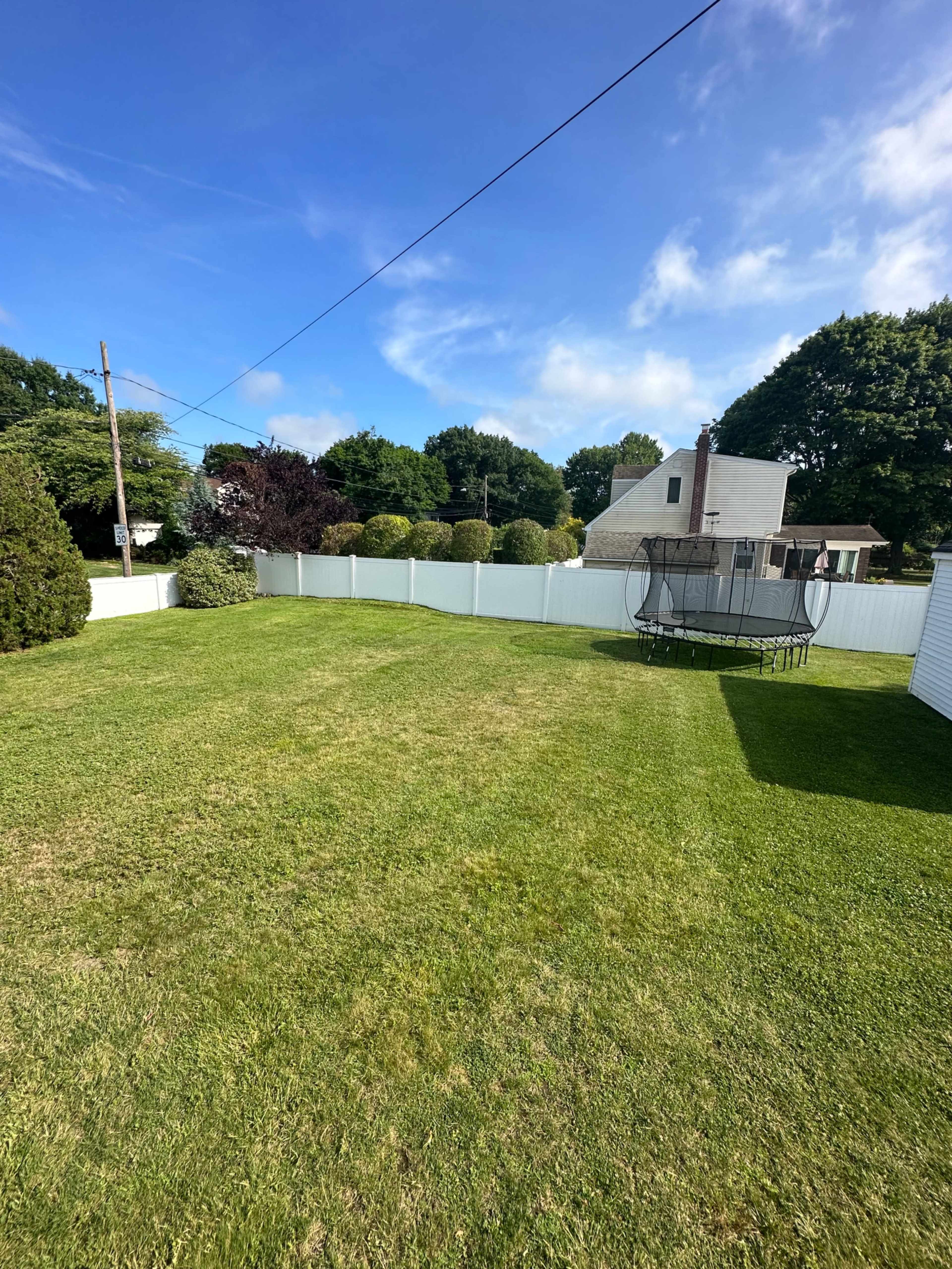 A spacious green backyard features a trampoline and a white fence, bordered by trees and houses in the background.