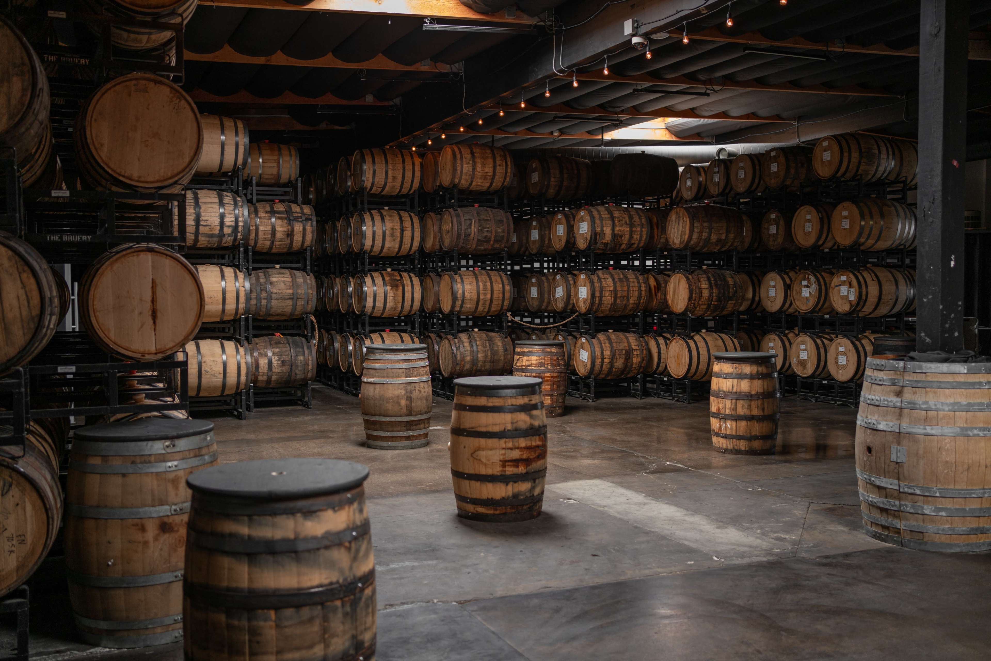 Rows of wooden barrels are stacked against the walls of a dimly lit venue, with additional barrels positioned on the floor.