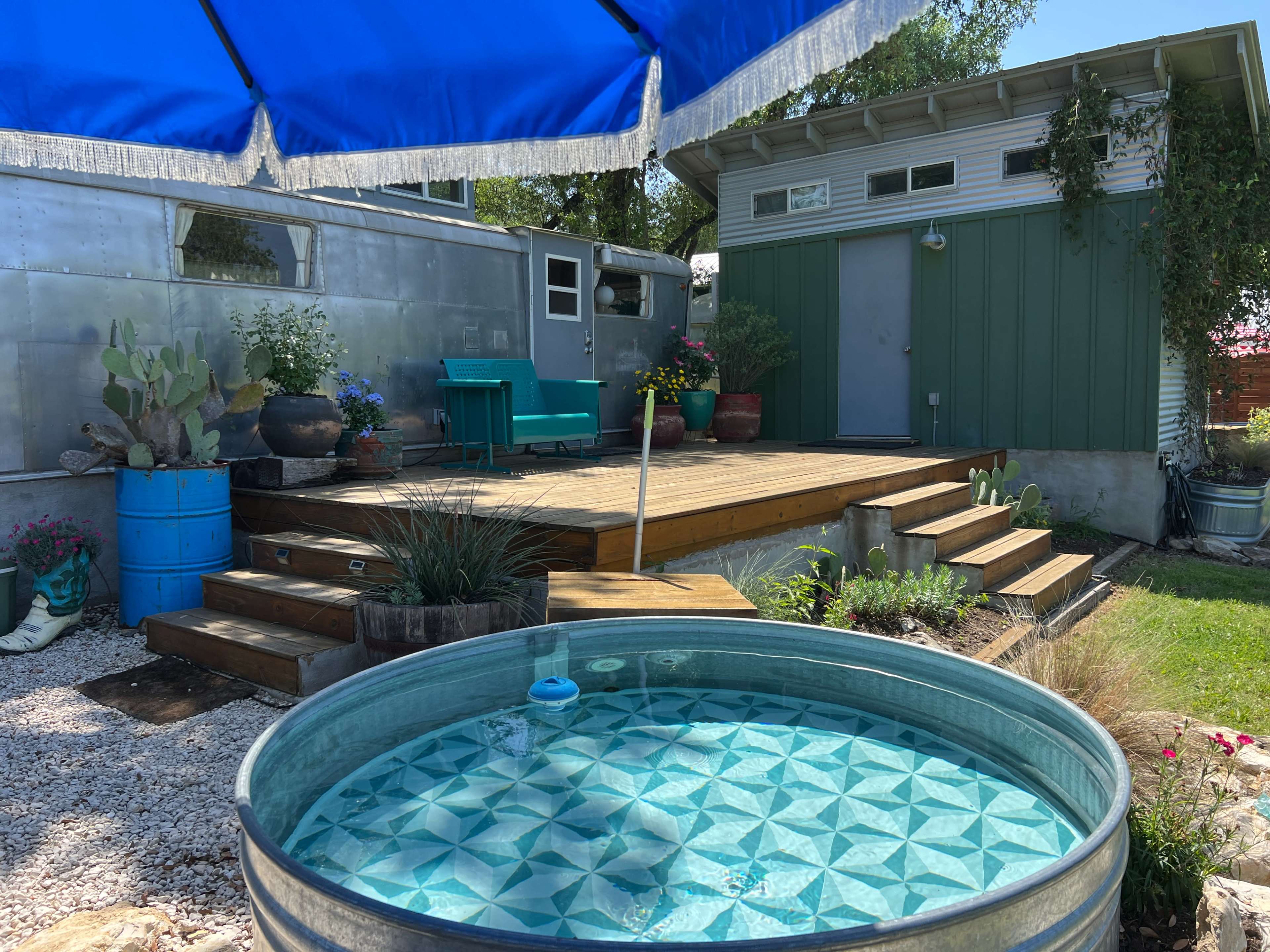 The image shows a backyard scene featuring a round metal water tank, a wooden deck with steps leading to a green shed, and a large blue umbrella providing shade.