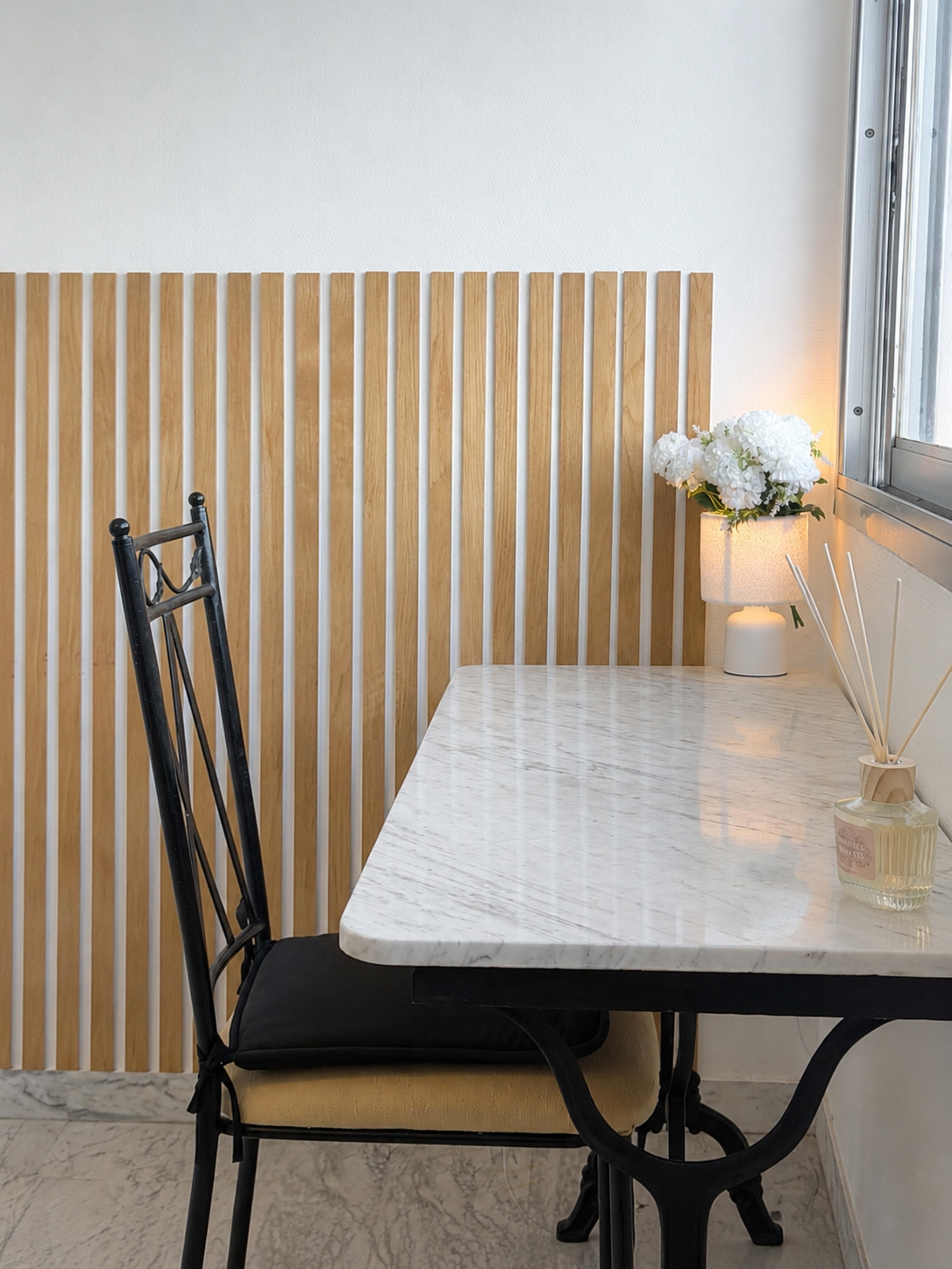 A marble table and a black chair are set against a wooden slatted wall, with a vase of flowers and a diffuser on the table.