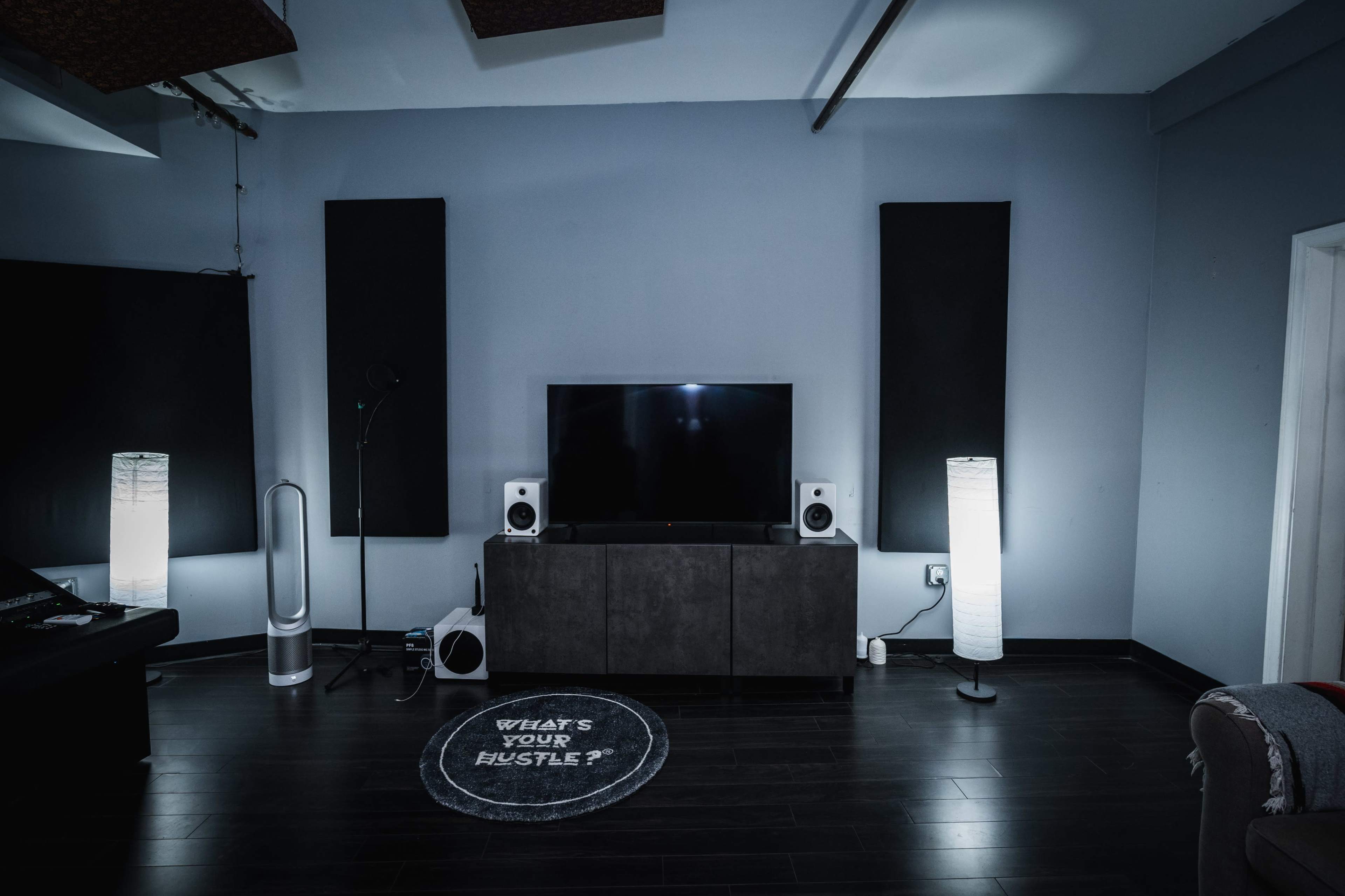 A modern interior space features a television mounted on a wall, flanked by speakers, illuminated by two floor lamps, with a circular rug displaying the phrase "WHAT'S YOUR HUSTLE?" on the floor.