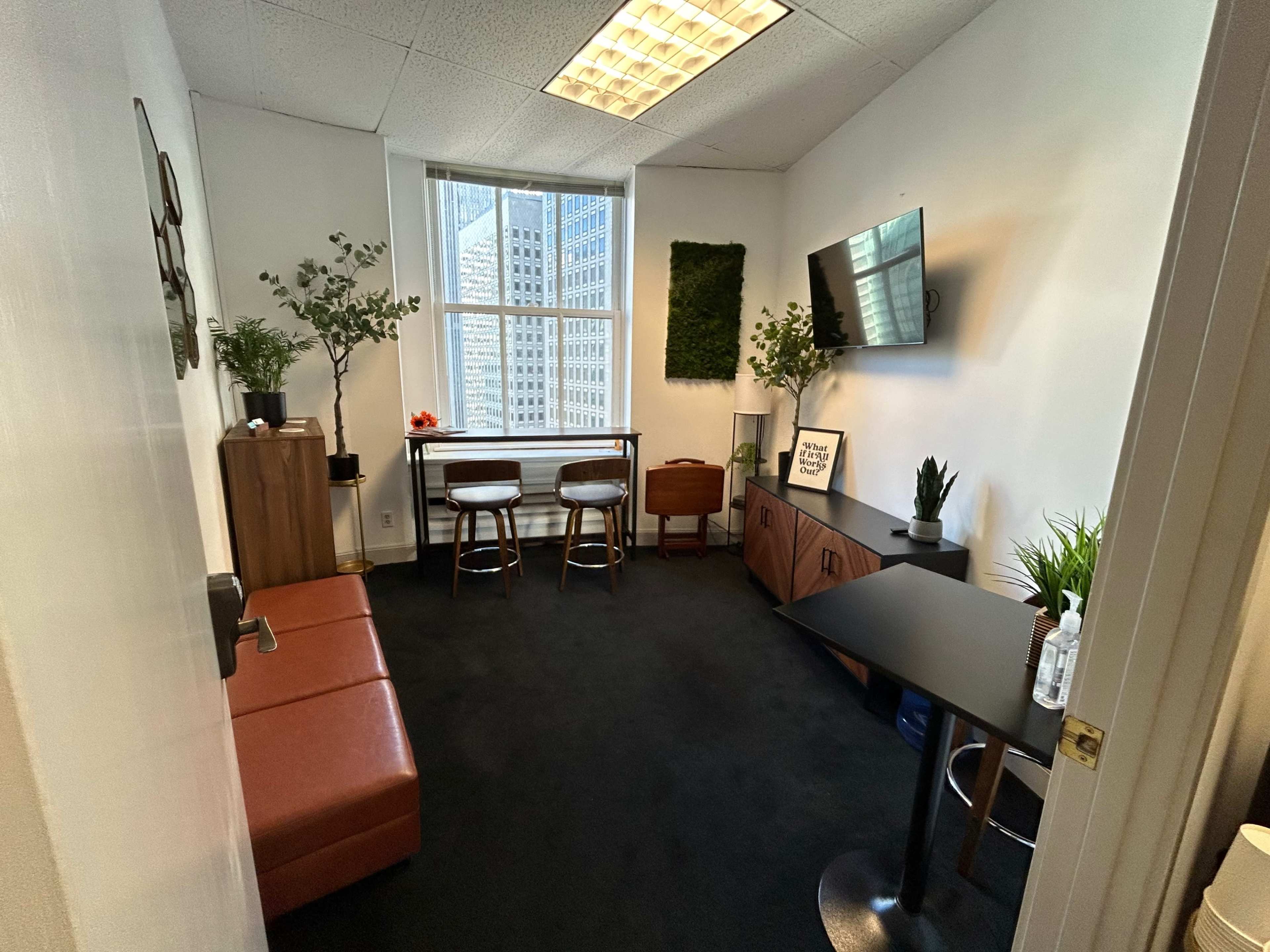 A small office space with a brown couch, two bar stools, a television mounted on the wall, and a few potted plants.