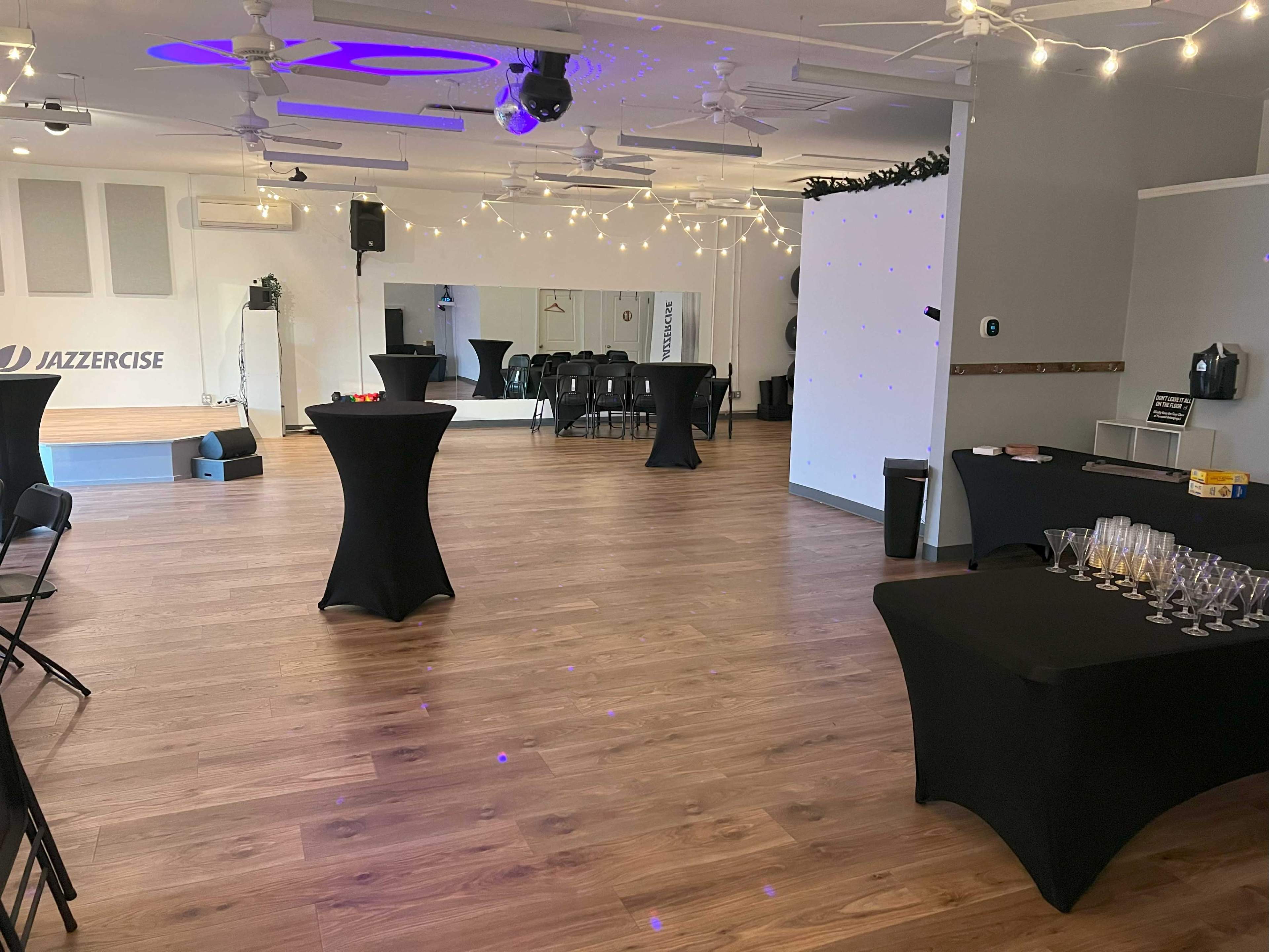 A spacious, well-lit event room with several black tables and chairs arranged for a gathering, featuring string lights overhead and a mirrored wall on one side.