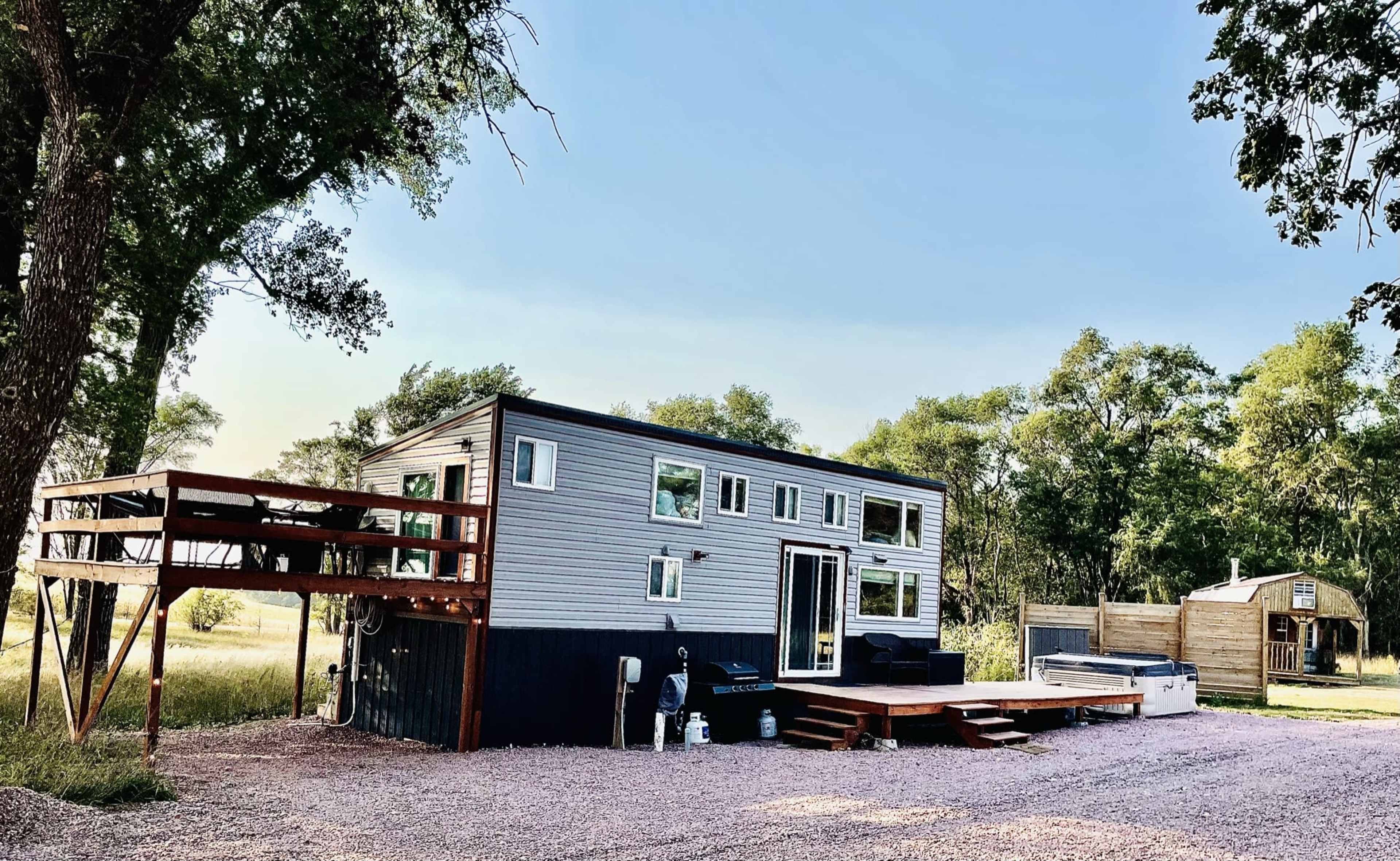 A modern tiny house with large windows and a deck sits on a gravel area surrounded by trees.