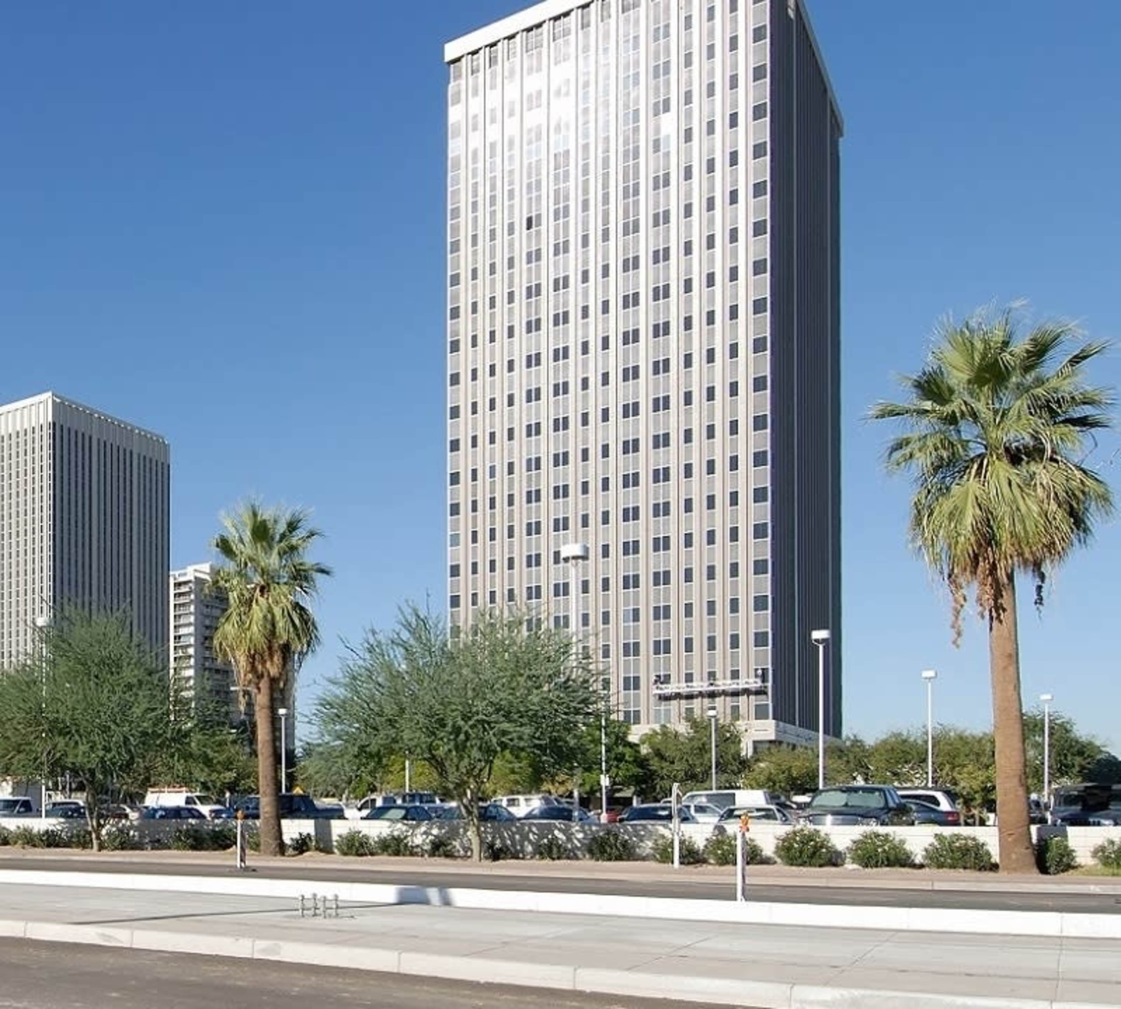 Two tall office buildings stand in a clear blue sky, surrounded by palm trees and parked cars in a landscaped area.