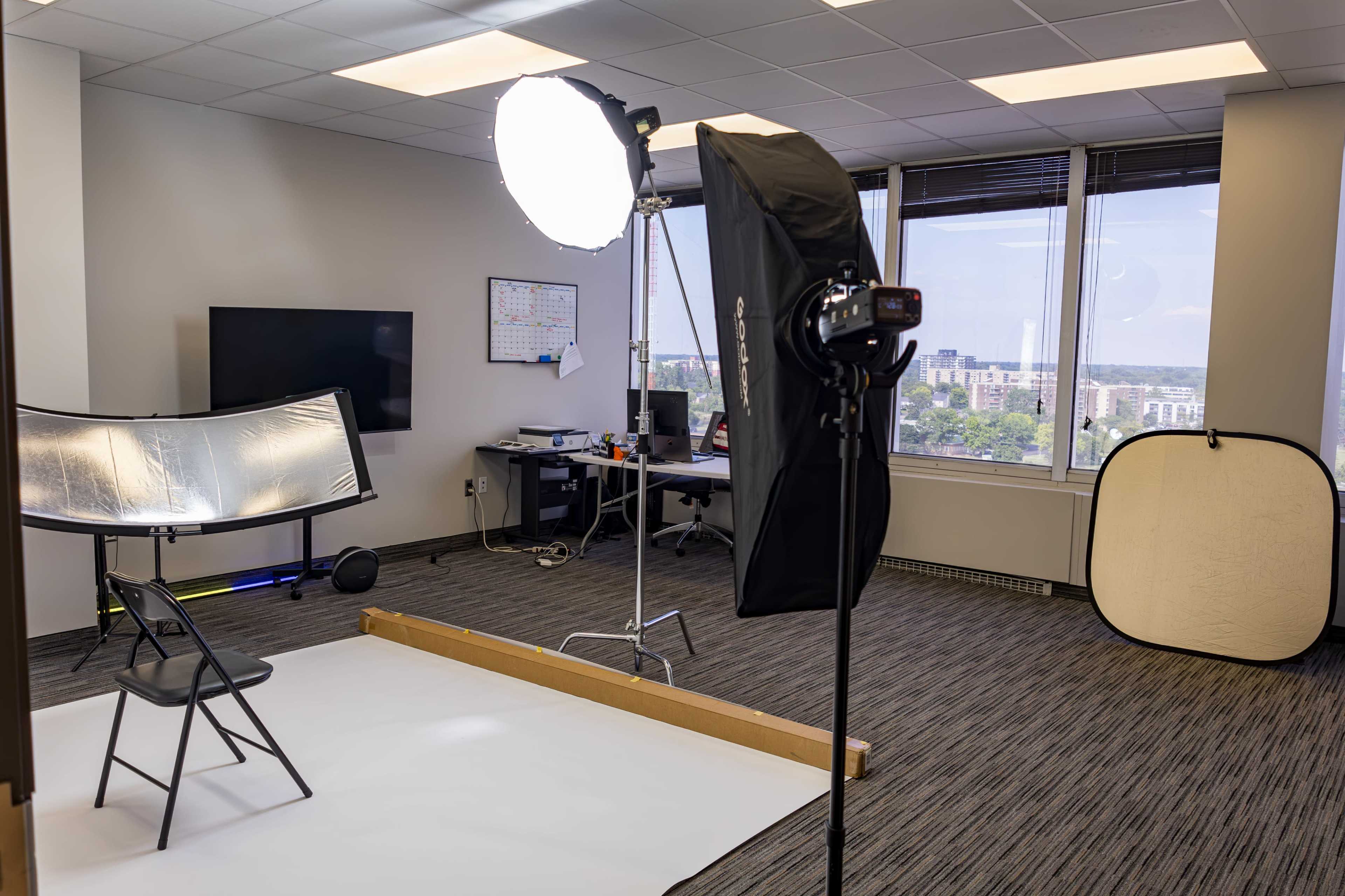 The image shows a photography studio setup in an office space, featuring a lighting setup with a softbox, a reflector, a chair, and a blank backdrop.