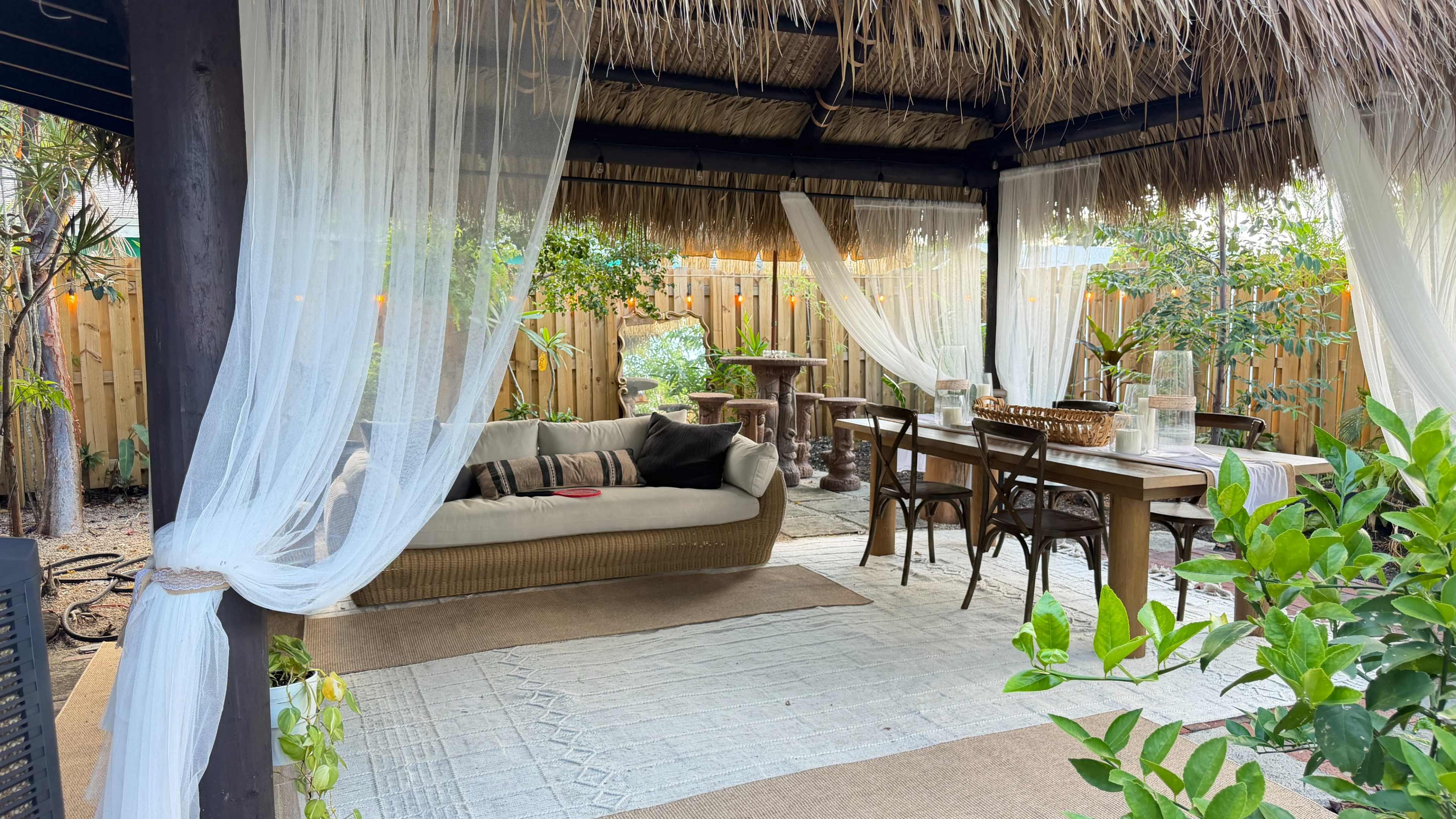 The image shows a shaded outdoor seating area with a thatched roof, featuring a sofa and a dining table surrounded by greenery.