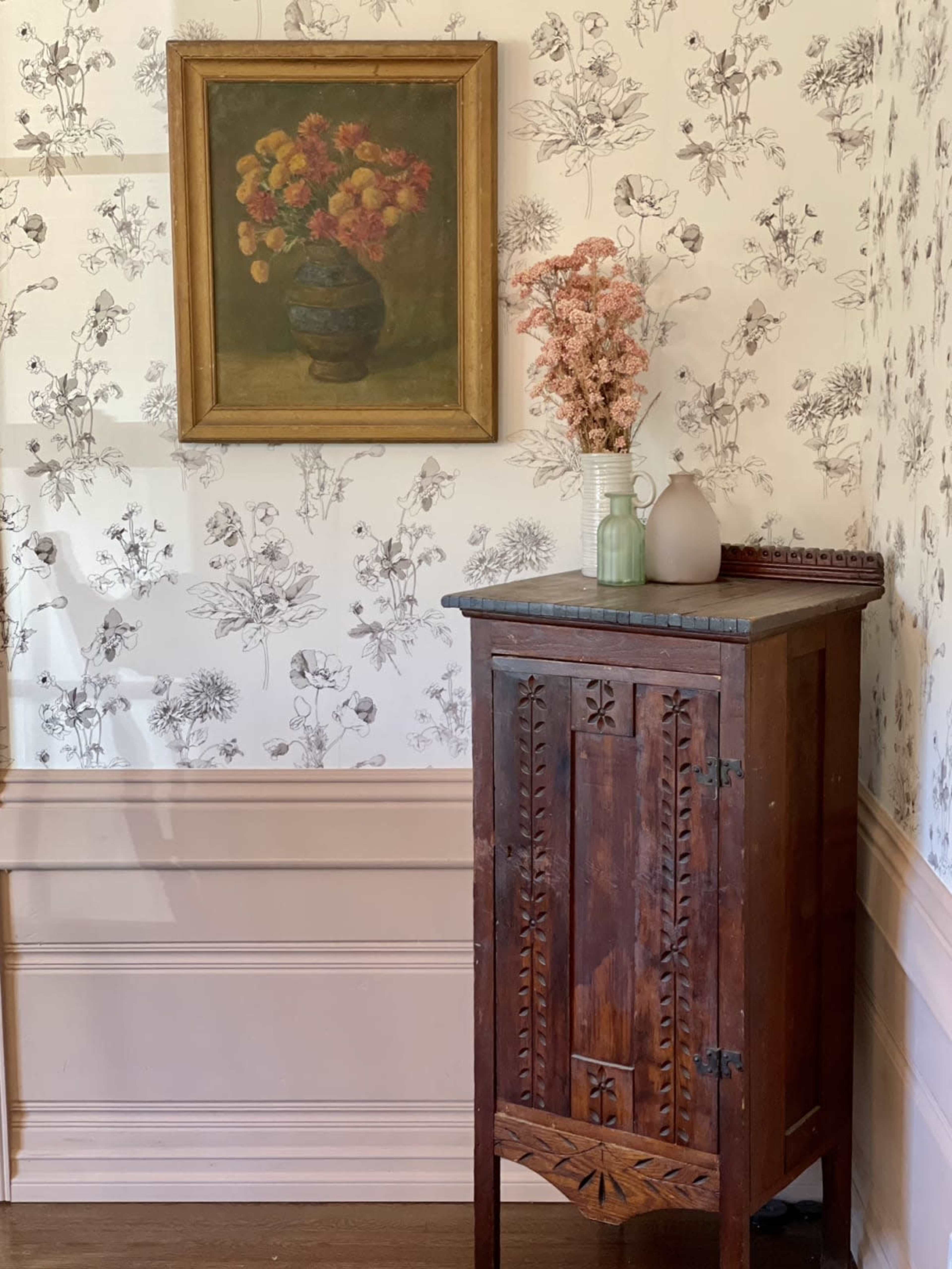 A wooden cabinet stands in a corner, displaying a vase of dried flowers and a framed painting of flowers on a patterned wallpaper background.