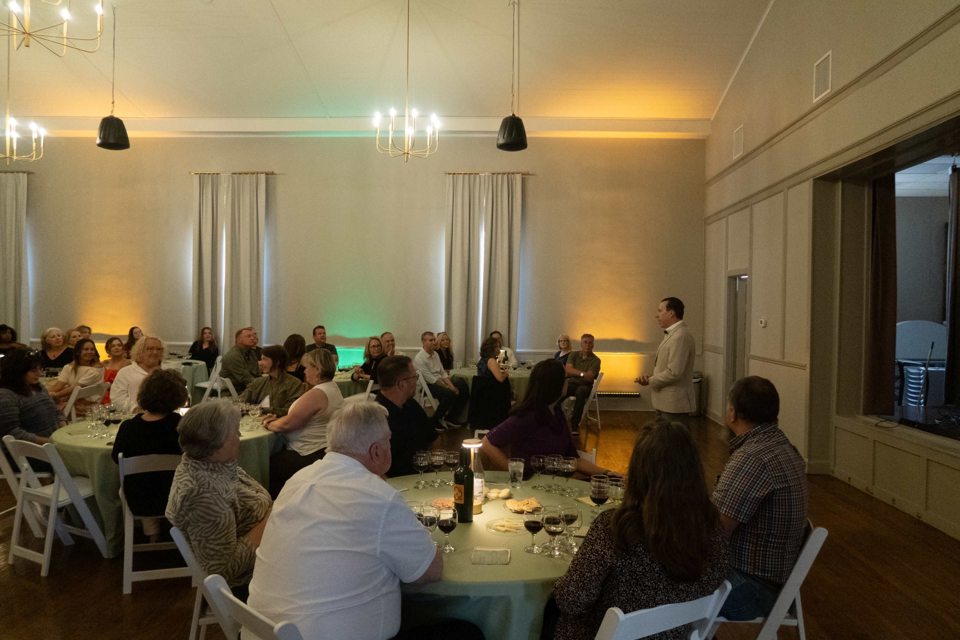 A speaker stands at the front of a well-lit room filled with seated attendees at round tables adorned with tablecloths and wine glasses.