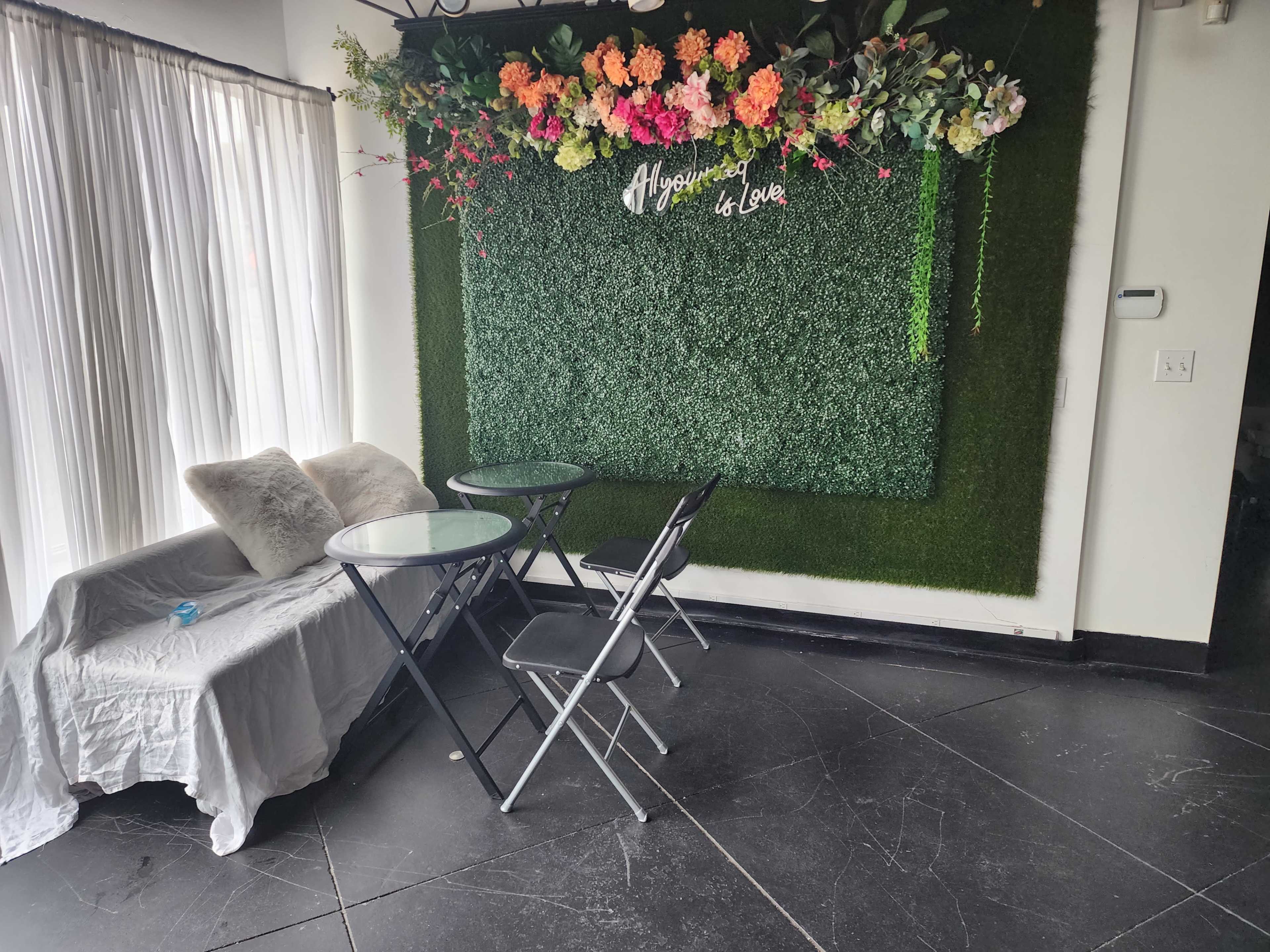 A small seating area with two black folding chairs and a glass table in front of an artificial green wall decorated with flowers, alongside sheer curtains.