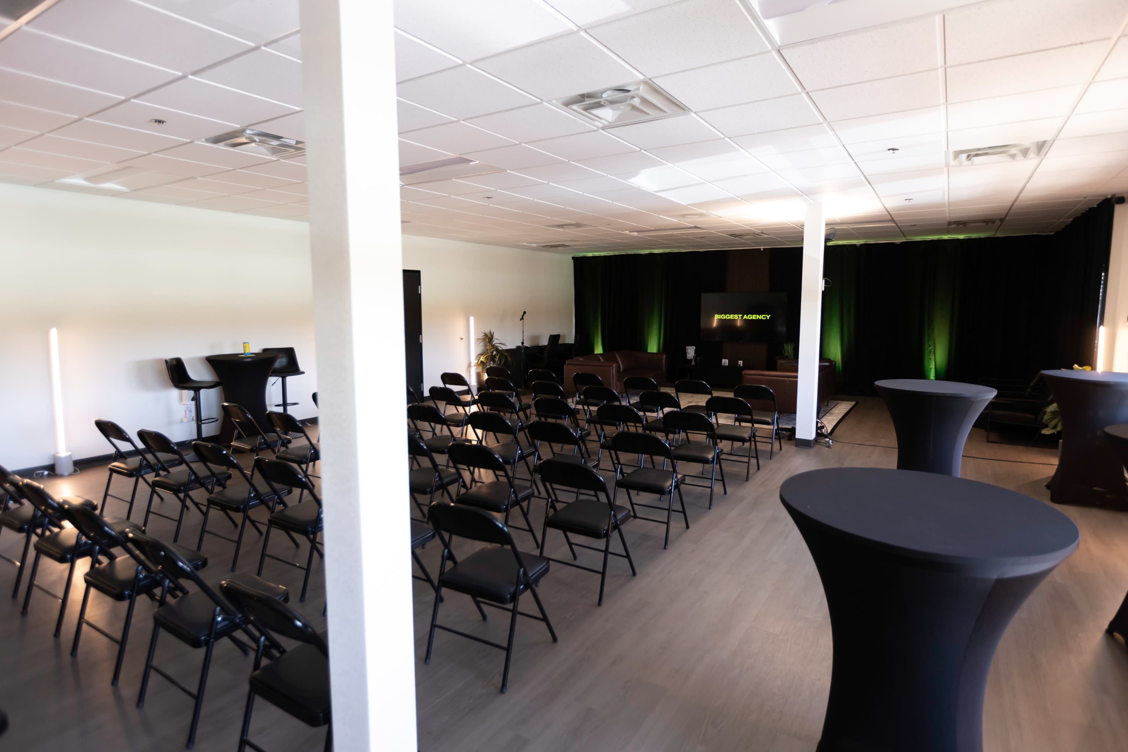 A spacious room with black chairs arranged in rows facing a stage, accompanied by high-top tables and green lighting.