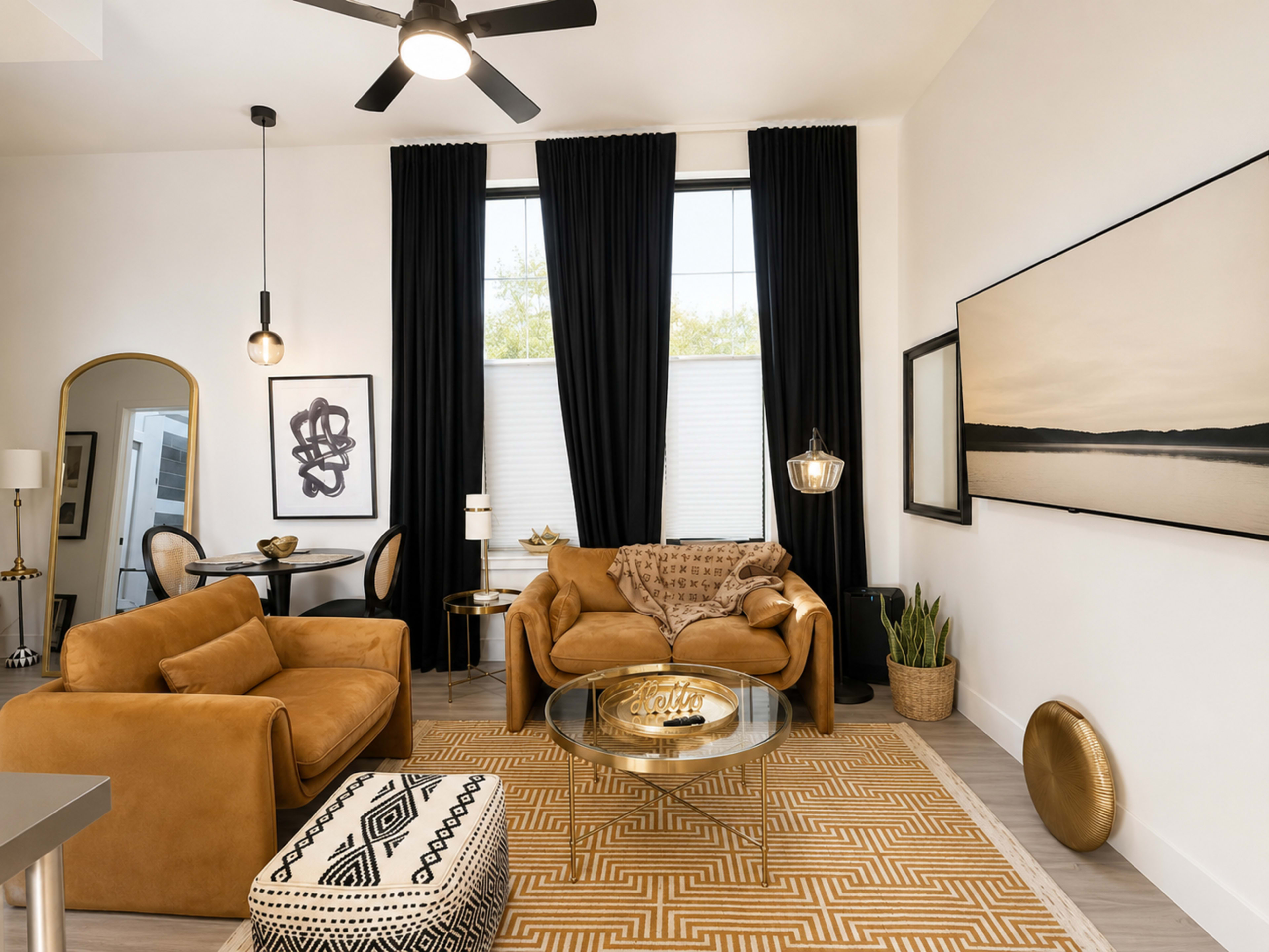 The image shows a modern living room with two brown couches, a round glass coffee table, and large windows covered by black curtains, all accented by decorative elements like artwork and plants.