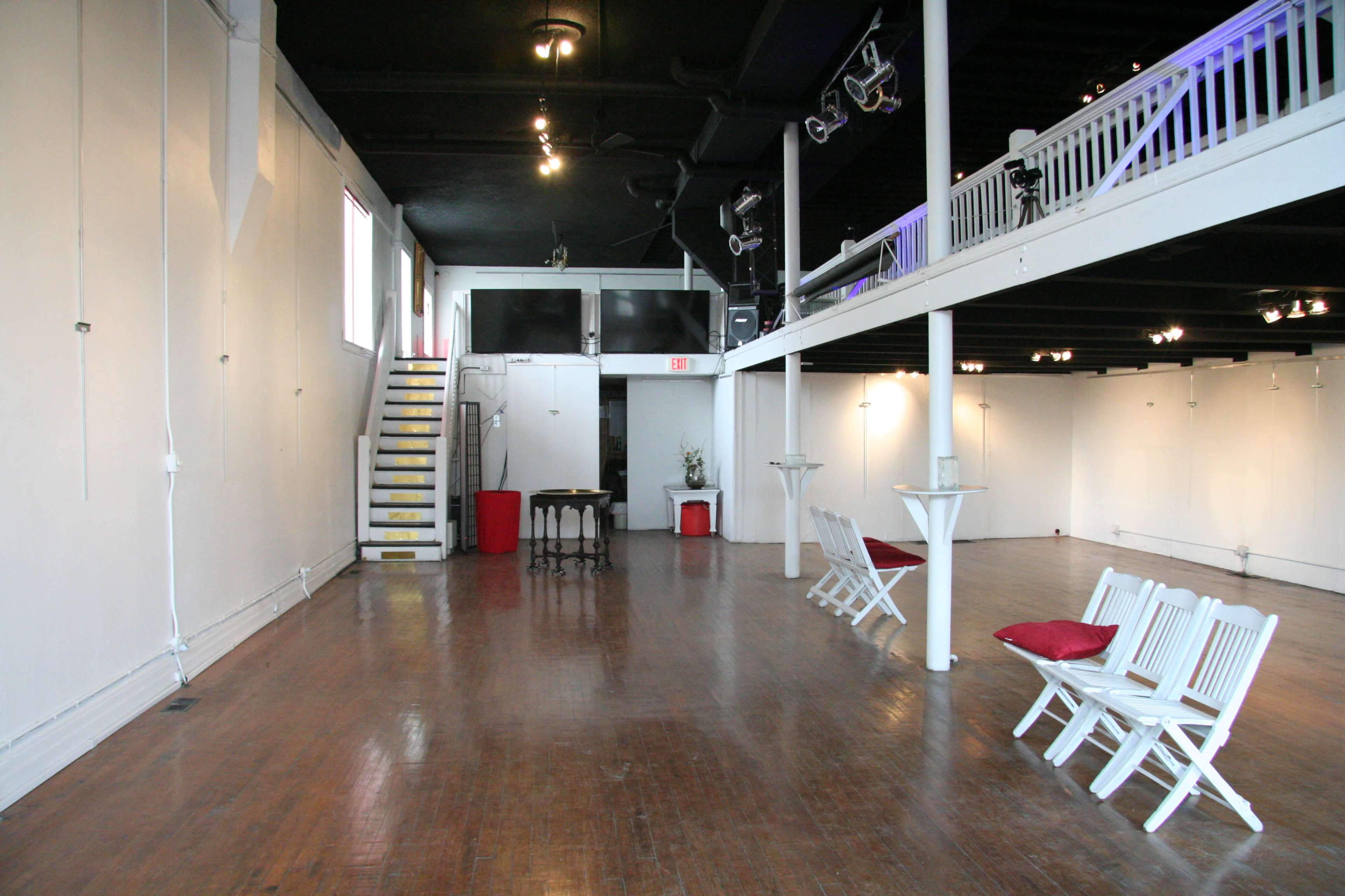A spacious, empty art gallery with wooden flooring, white walls, a staircase, and a few empty chairs arranged around small tables.